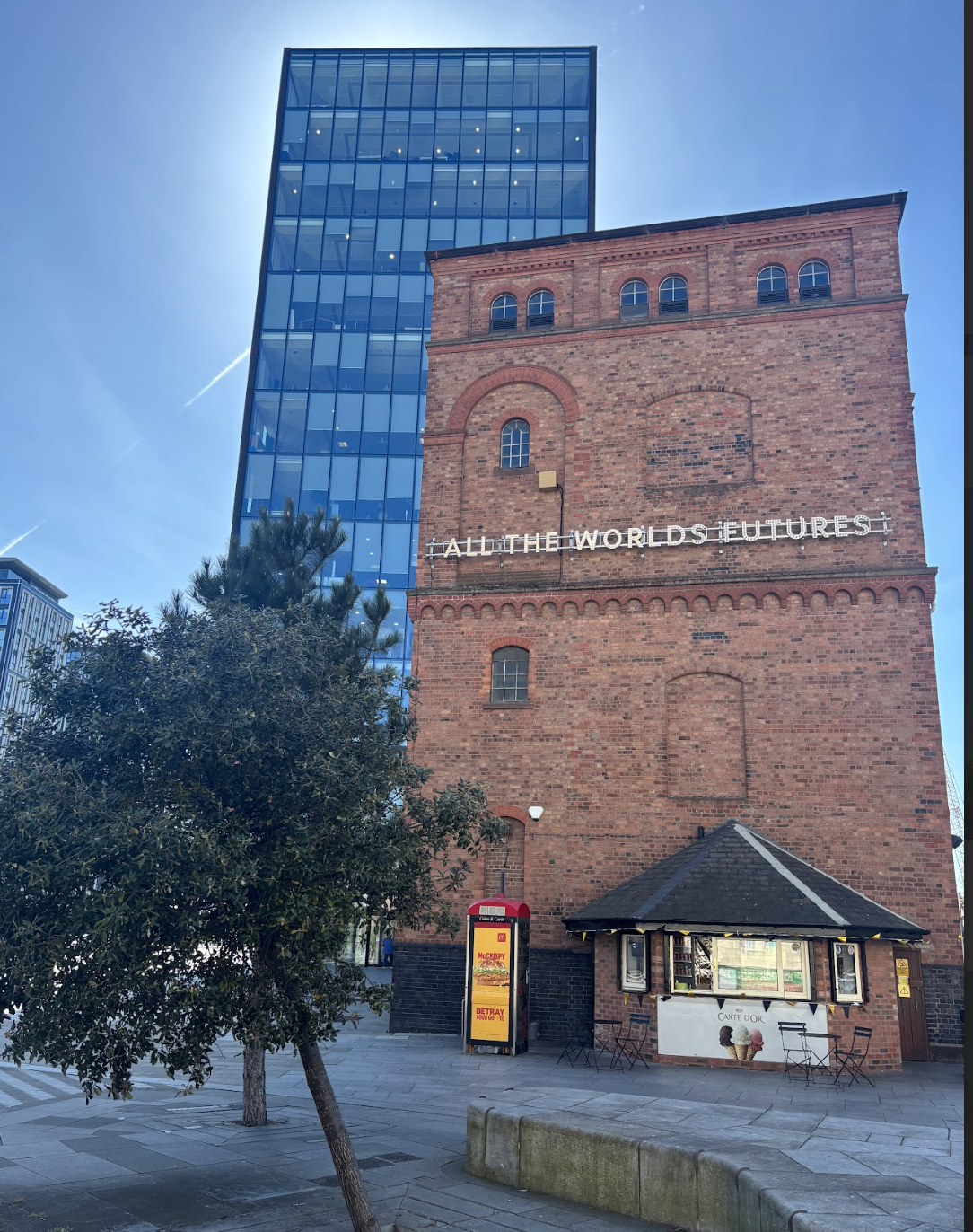 A tall, modern glass building beside an older red brick building with a sign that says 'ALL THE WORLD'S FUTURES.' There is a small kiosk and outdoor seating area in front of the brick building, with a tree partially obscuring the view.