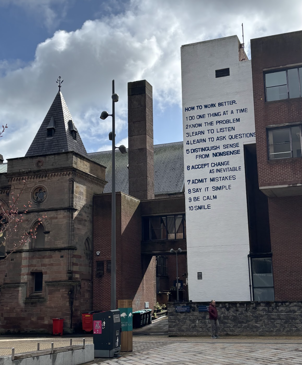 A cityscape with old and modern buildings. A large white wall features a list titled 'HOW TO WORK BETTER,' with 10 numbered points. A person stands near the wall, and there are trees with pink blossoms.