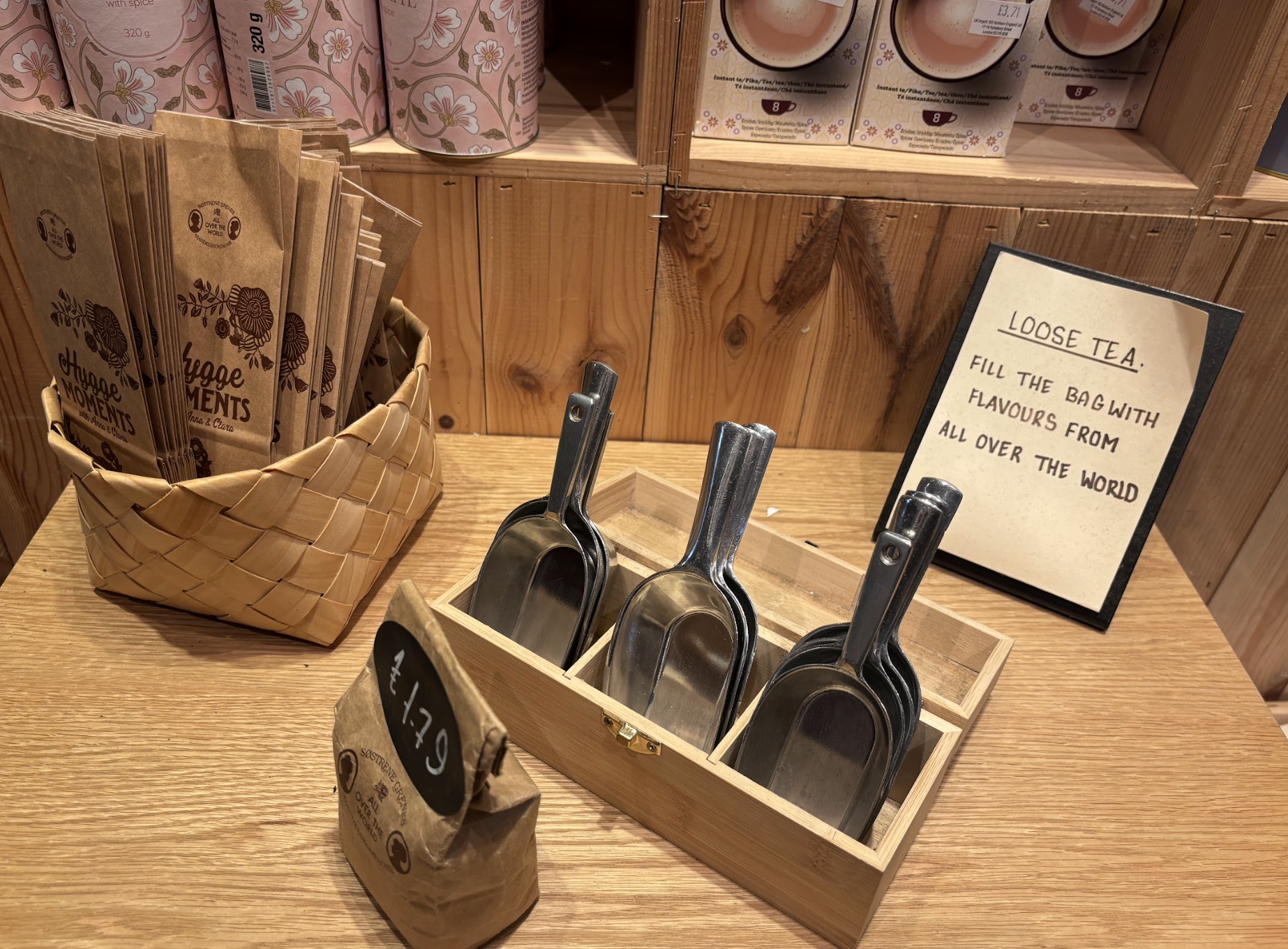 Display of various loose tea options in a store, including a basket of tea bags labeled "Loose Moments," a small wooden box with metal scoops, and a handwritten sign encouraging customers to fill bags with flavors from around the world.