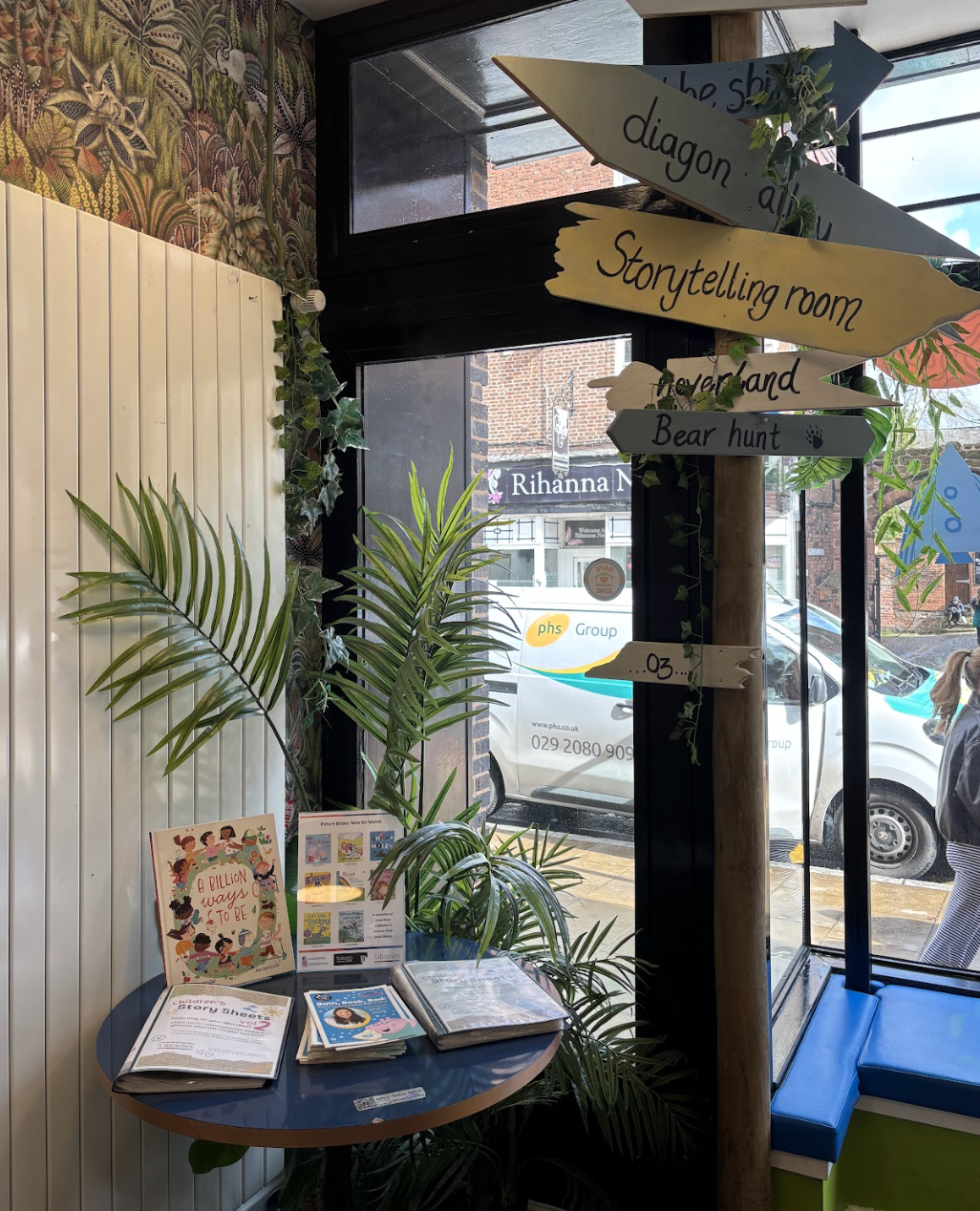 Interior of a room with a blue table holding children's books and above, a decorative wooden signpost with colorful arrows indicating various themed rooms such as 'storytelling room' and 'bear hunt.' There are green plants near the window and a view of parked cars outside.