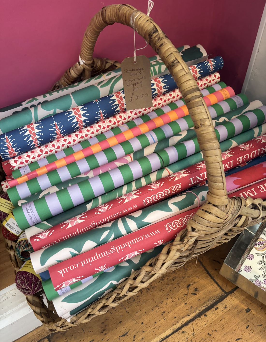 A wicker basket filled with rolls of Christmas wrapping paper in various festive patterns and colors, with a wooden handle and a handwritten price tag.