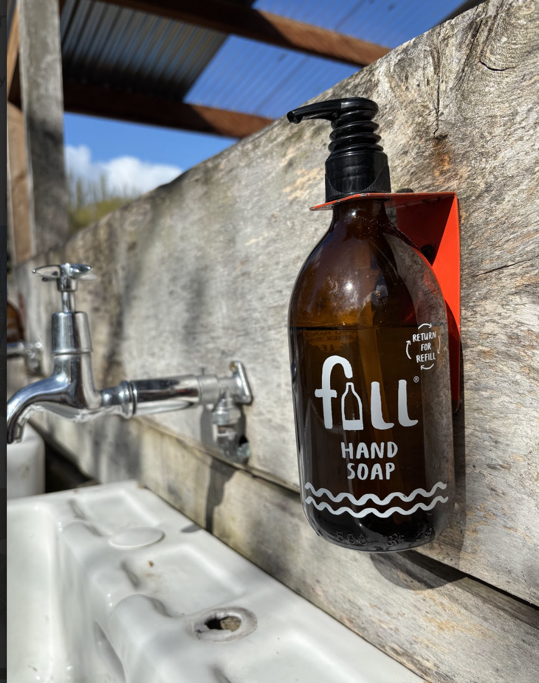 A brown glass bottle with a black pump dispenser labeled "fall hand soap" mounted on a rustic wooden wall, with a white sink and silver faucet in the background.