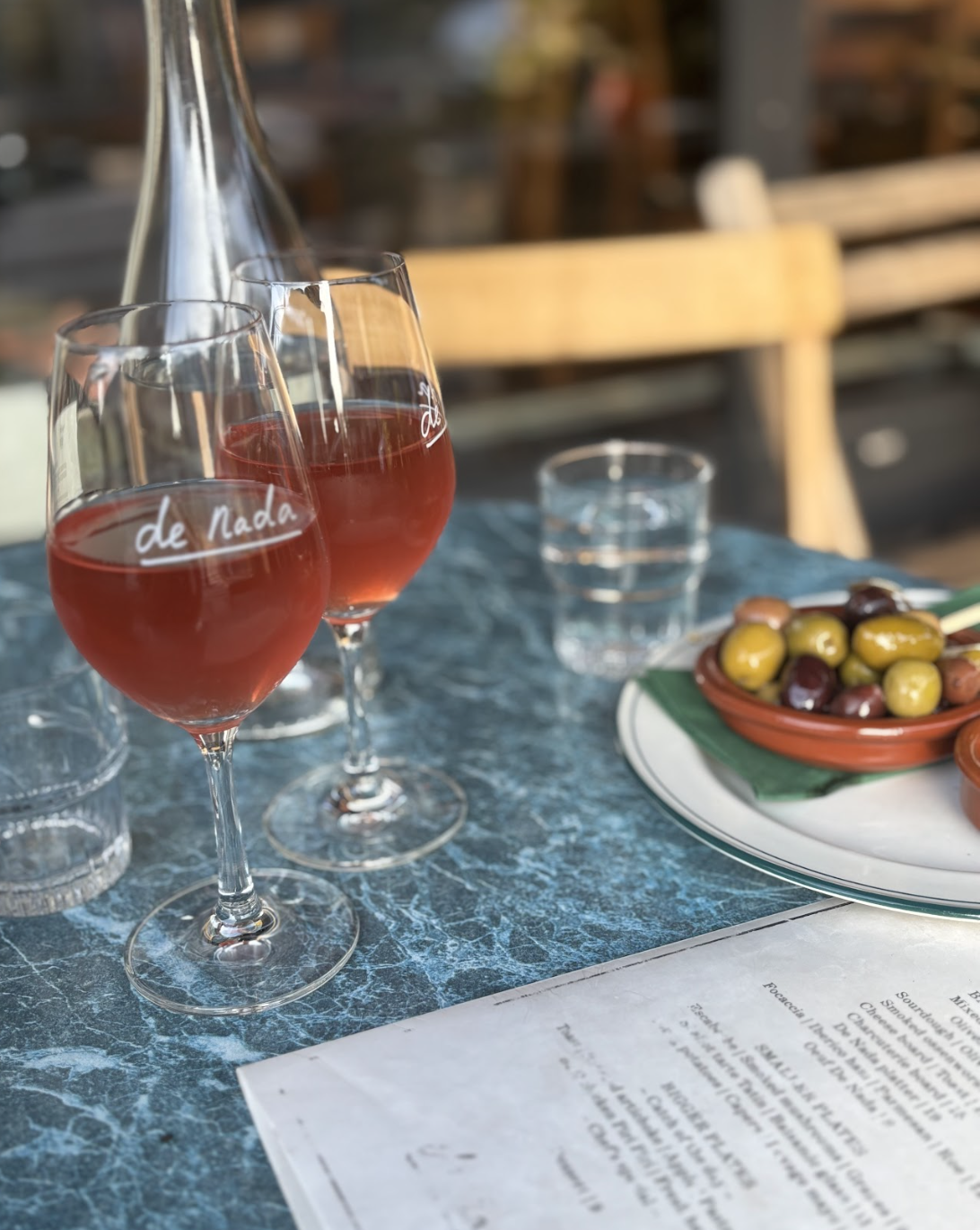 Two glasses of rosé wine on a blue marble table with a pitcher behind them, a clear glass of water, a bowl of olives, and a partially visible menu.