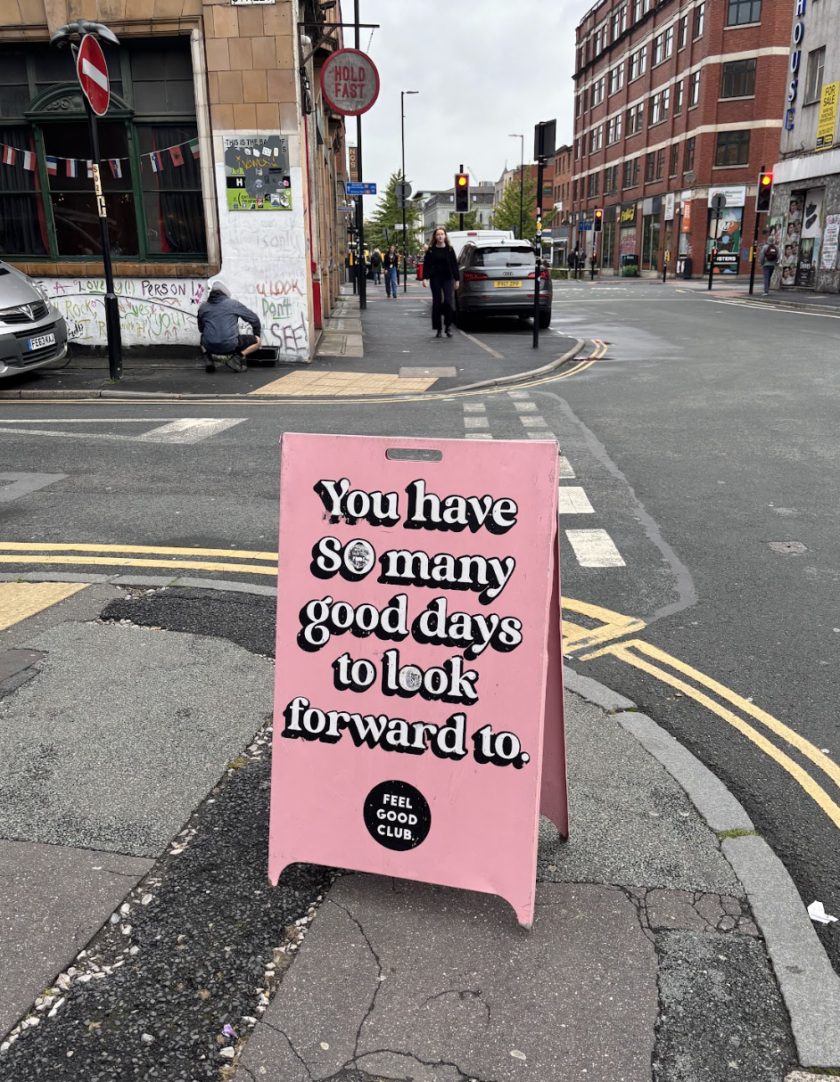 A pink sidewalk sign on a city street reads 'You have so many good days to look forward to.' in large black and white text with a small black circle at the bottom that says 'FEEL GOOD CLUB.' In the background, there are traffic lights, cars, pedestrians, a building with graffiti, and a person squatting near the graffiti wall.