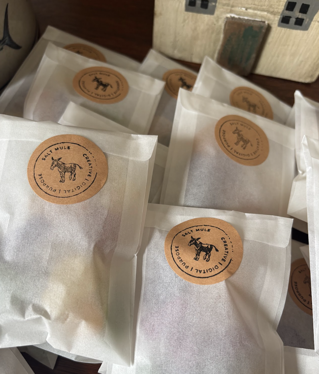 Several white paper bags with orange circular labels featuring a black outline drawing of a mule, the words 'Salt Mule' and 'Cognitive Disorder,' and the phrase 'Creative.' The bags are arranged on a flat surface.