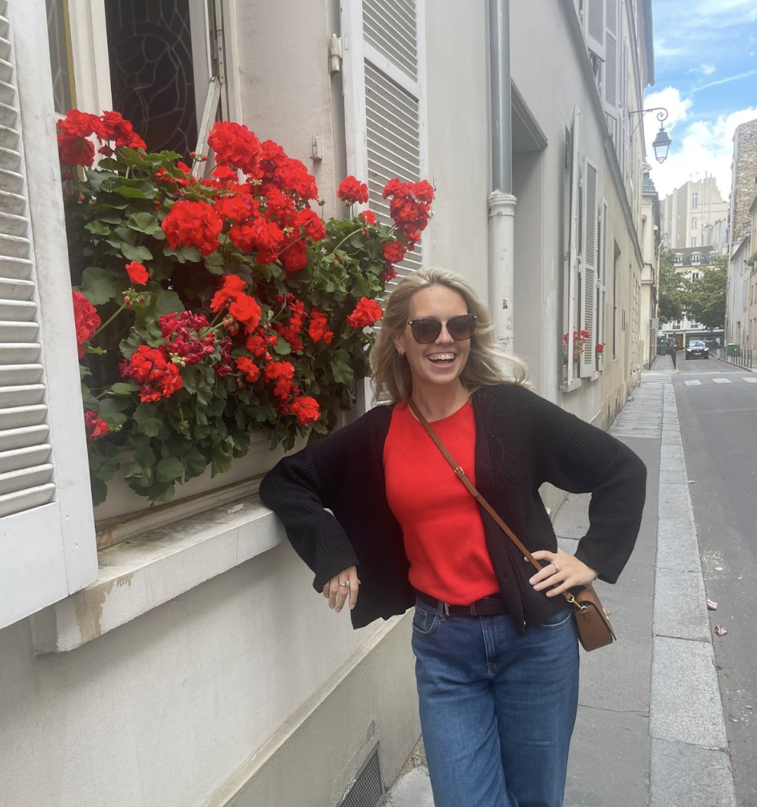 A woman smiling and wearing sunglasses, a red shirt, and a black cardigan, standing on a city street next to a window with red flowers in a window box.