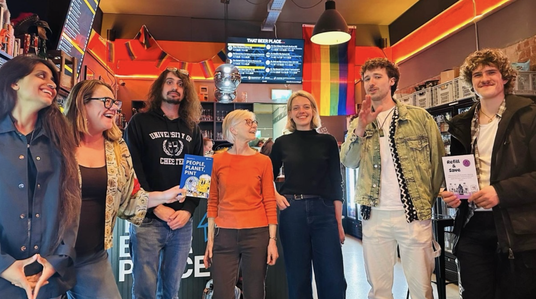 A group of seven diverse people standing inside a cafe or bar, smiling and talking. They are holding flyers about environmental issues and beverage recycling. The background features a rainbow pride flag, a menu, and a TV screen. The atmosphere is casual and friendly.