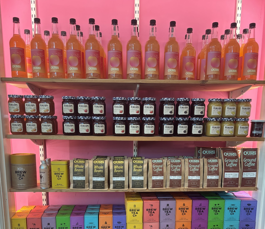 Shelf with bottles of peach-flavored drink, jars of jam or jelly, bags of whole beans and ground coffee, and boxes of tea in a grocery store.