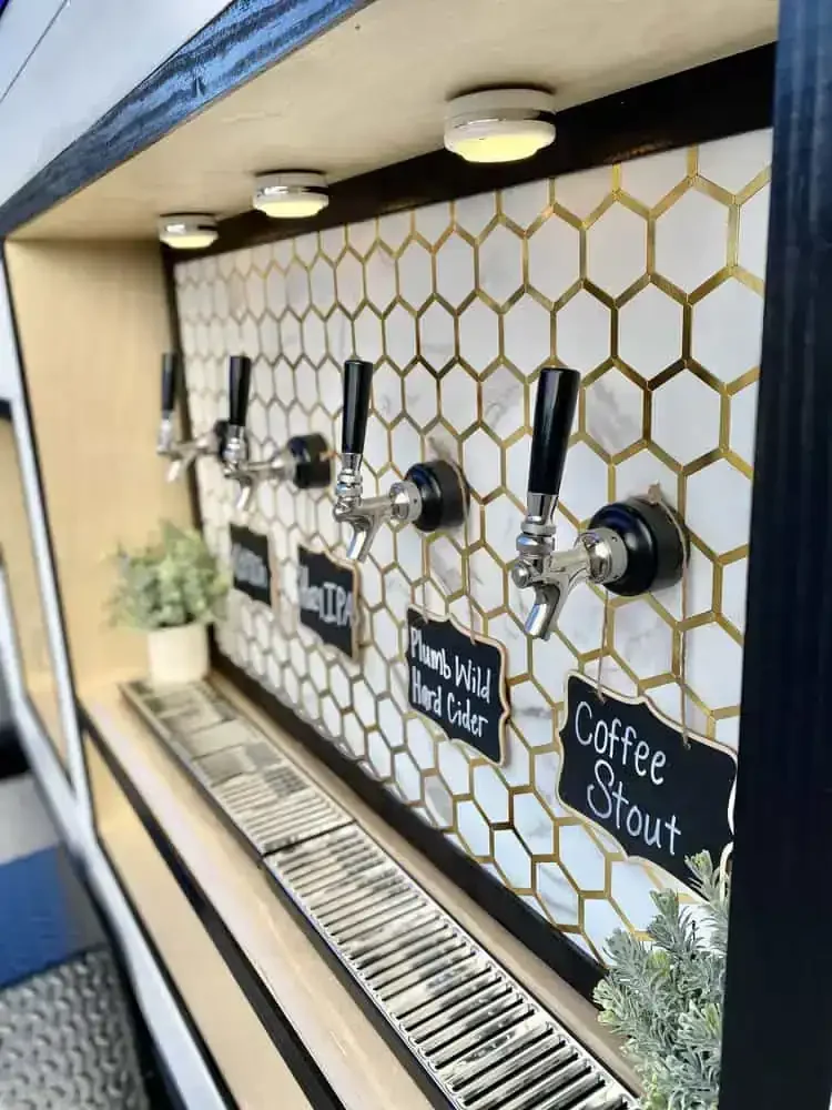Four beer taps labeled IPA, Plumb Wild Hard Cider, and Coffee Stout at a bar counter with a honeycomb-patterned background and small chalkboard signs.