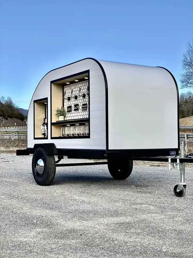 A mobile bar trailer with an outdoor serving window, shelves with glasses and bottles, and minimalistic decor, set against a clear blue sky.