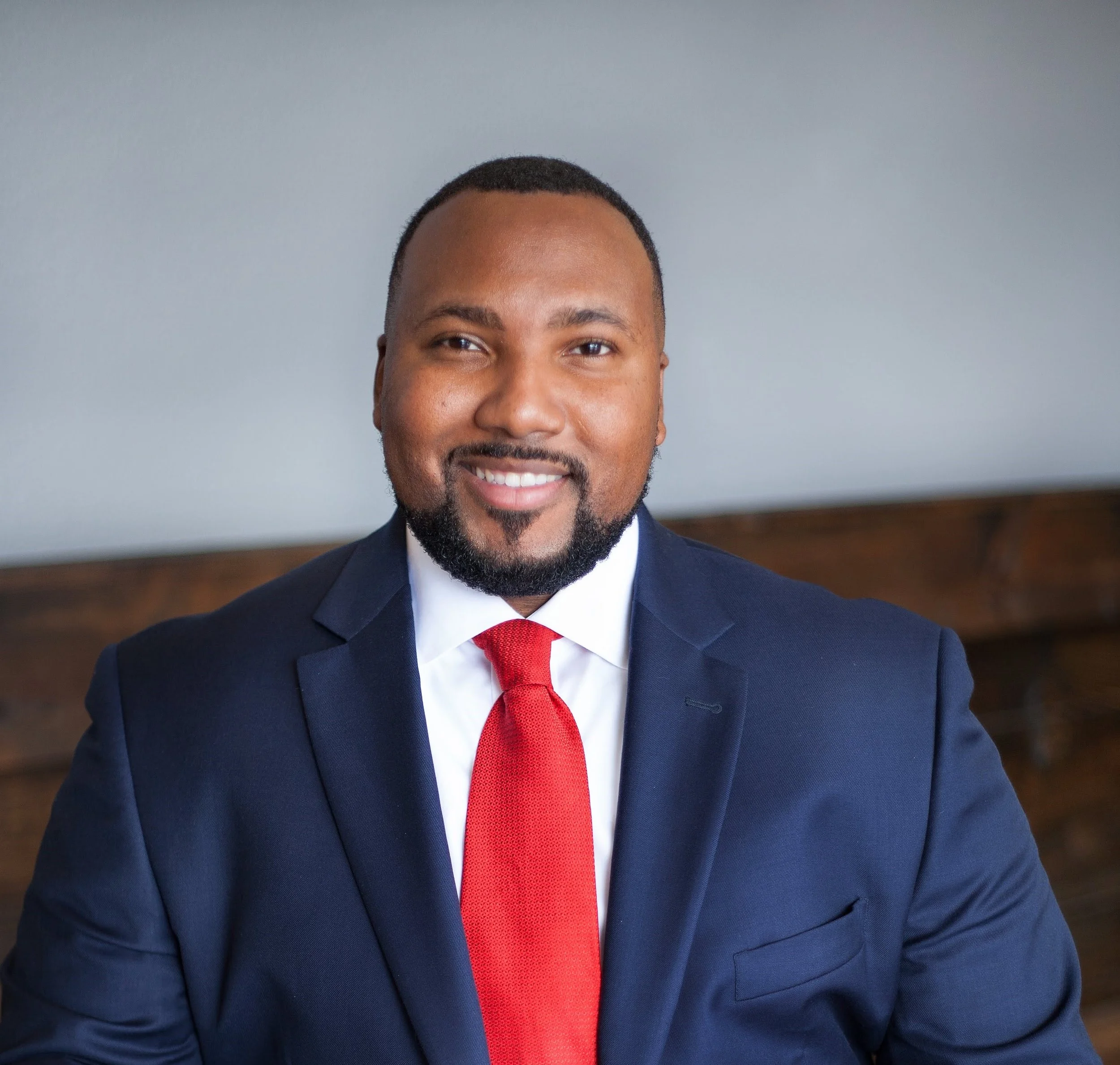 A professional man in a navy suit, white shirt, and red tie smiling for a portrait inside a room with a dark wood feature in the background.
