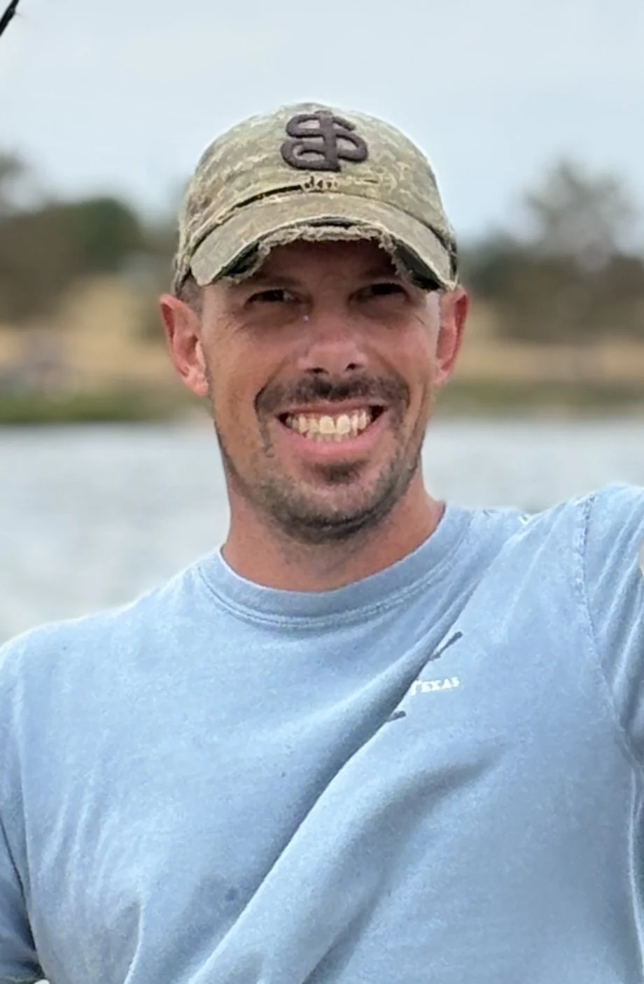 A man smiling outdoors, wearing a camouflage baseball cap and a light blue t-shirt with a small white logo on the chest, standing near a body of water with trees in the background.