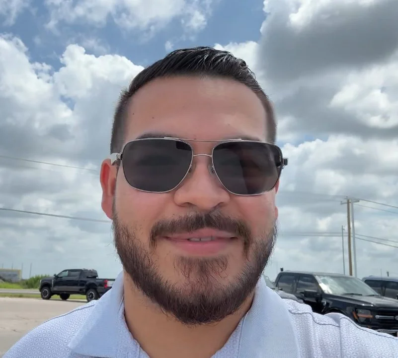 A man with dark hair, beard, and sunglasses smiling outdoors on a partly cloudy day, with cars and power lines in the background.