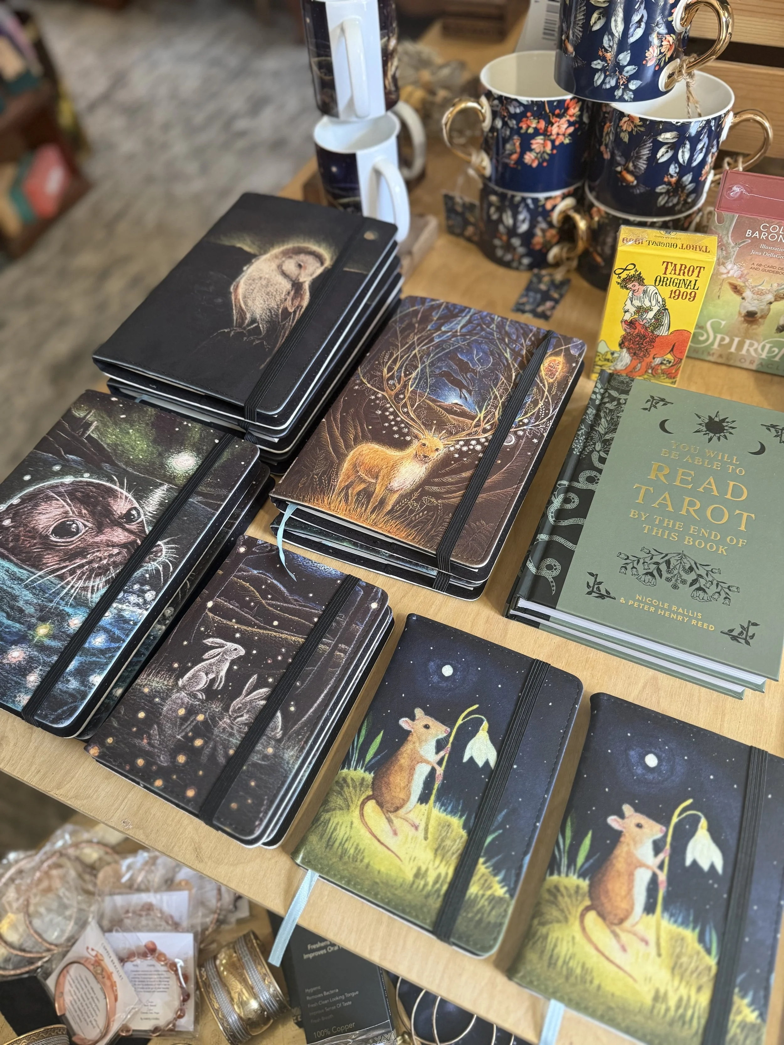 Arrange of notebooks with animal and nature illustrations, including a deer, a fox, and a bear, on a wooden table with cups and tarot cards.