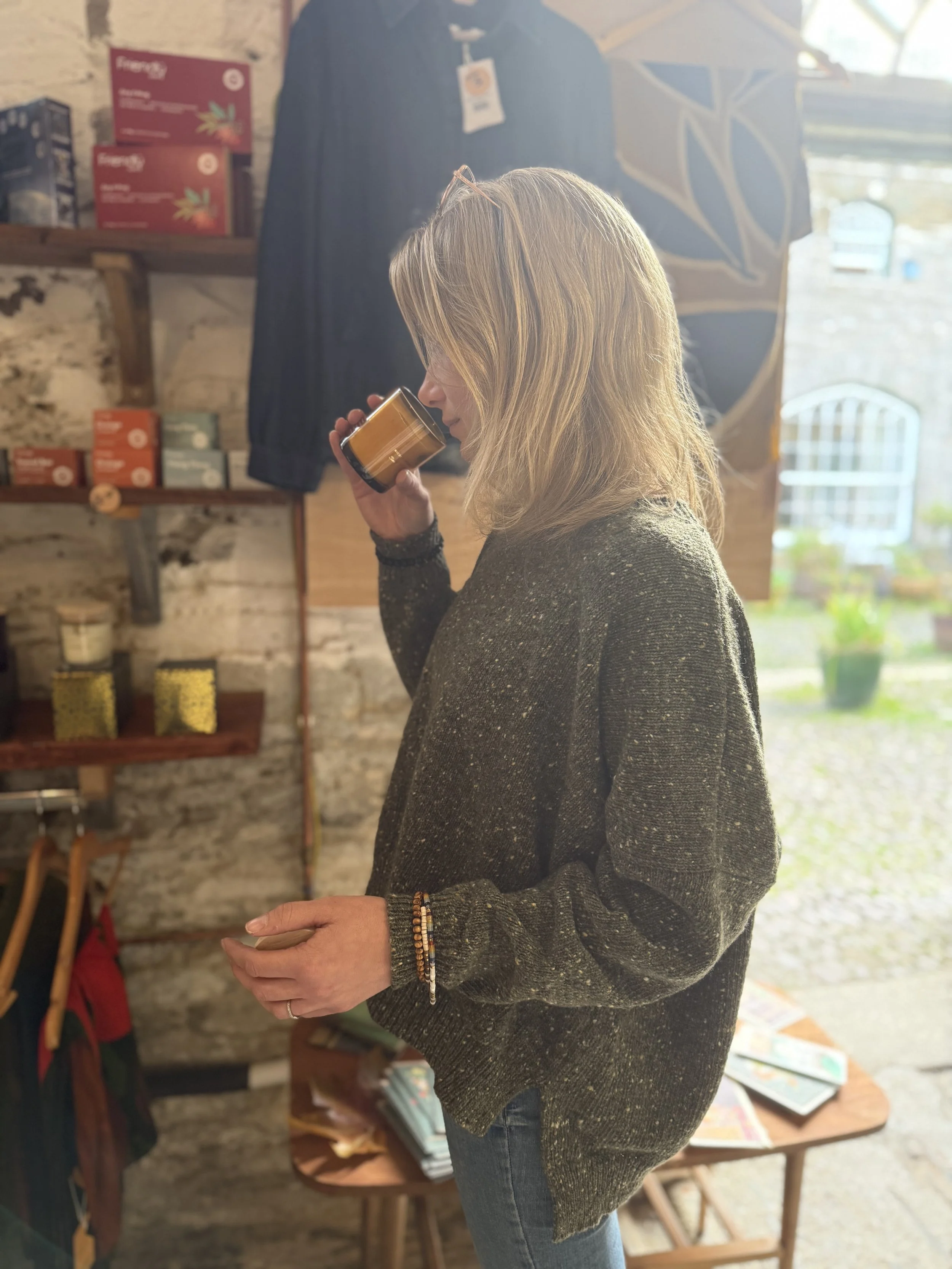 Woman with blonde hair holding a candle in a shop with wooden shelves and colorful boxes, wearing a dark sweater and beaded bracelets.