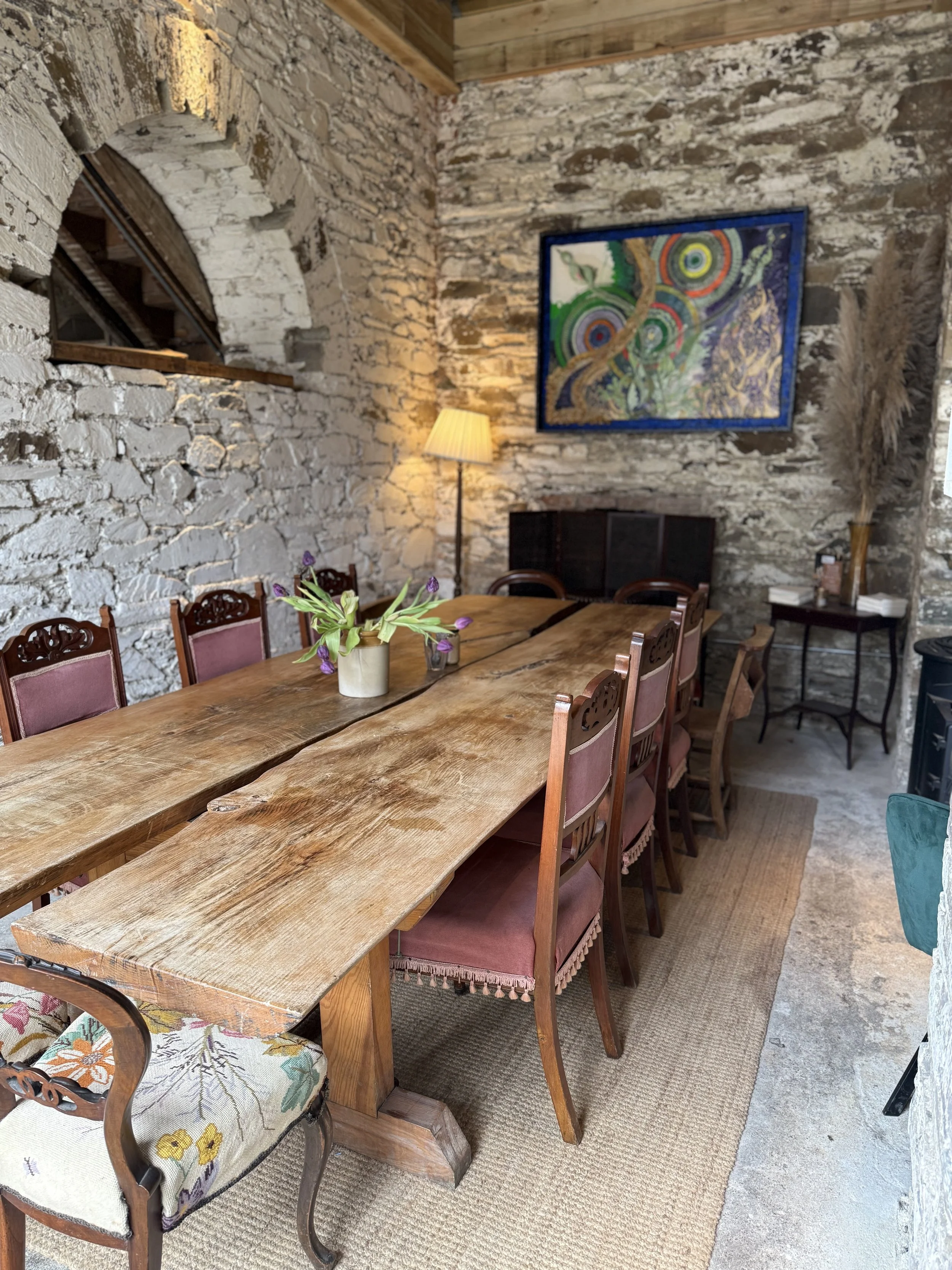 A dining area with a long wooden table surrounded by chairs with pink upholstery and decorative cushions. There is a floral centerpiece on the table, a lamp on the back wall, and a colorful abstract painting on the stone wall. The room has exposed st