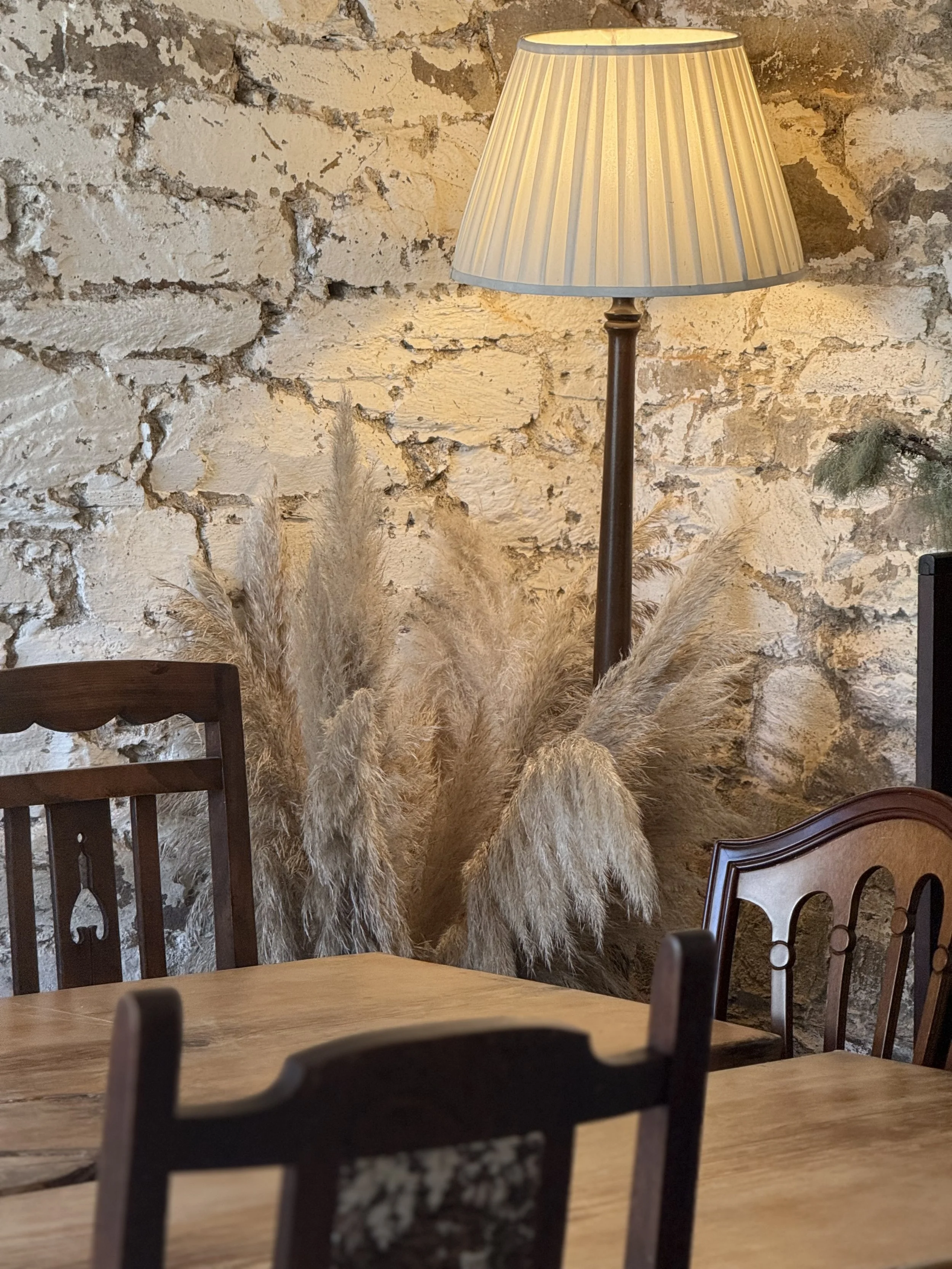 Interior photo of a rustic dining room with a wooden table, wooden chairs, a stone wall, a floor lamp with a beige lampshade, and a decorative arrangement of dried pampas grass.