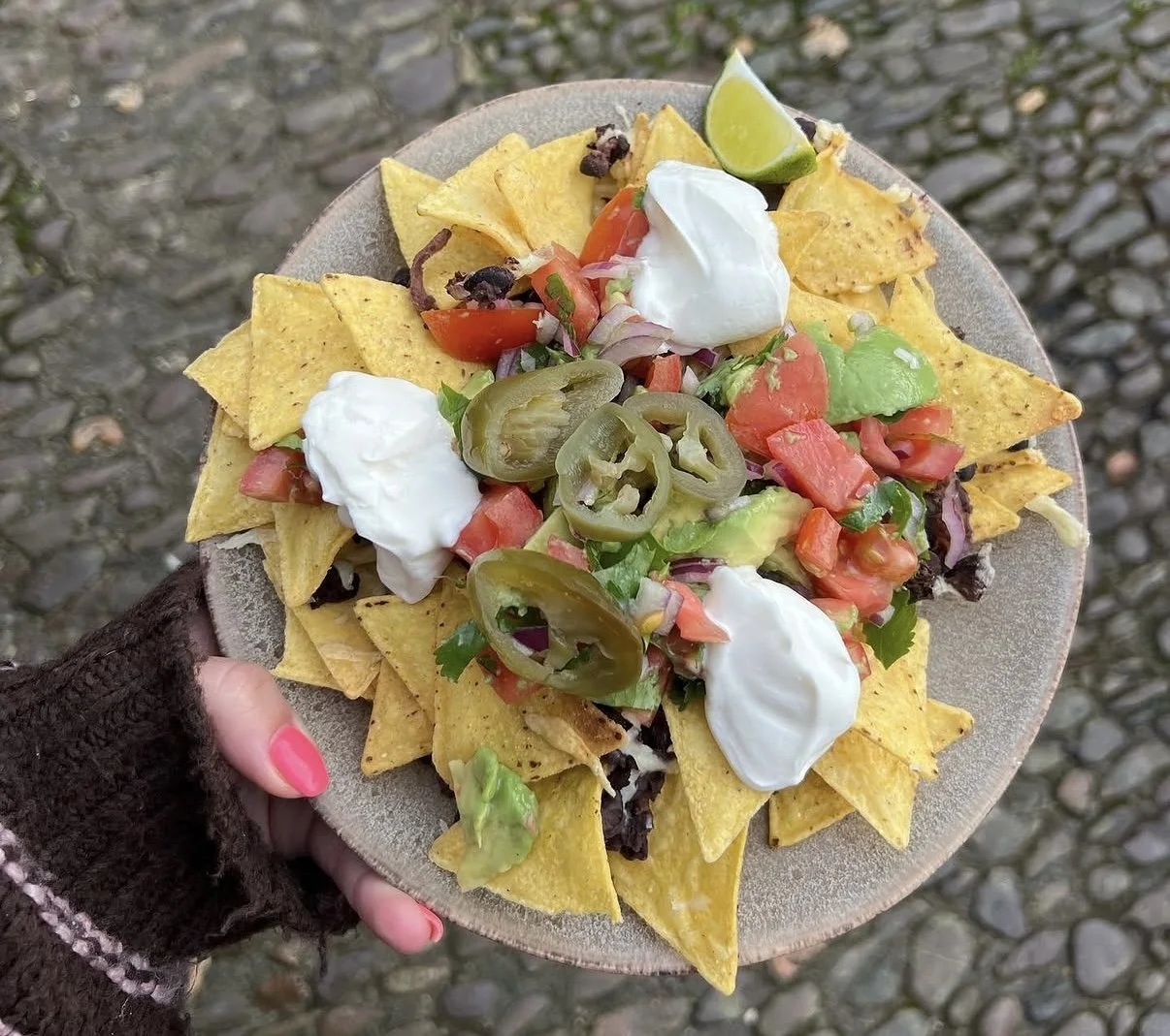 A plate of nachos topped with sour cream, jalapeños, diced tomatoes, chopped onions, shredded lettuce, black beans, and a lime wedge, held by a person with pink nail polish against a cobblestone background.