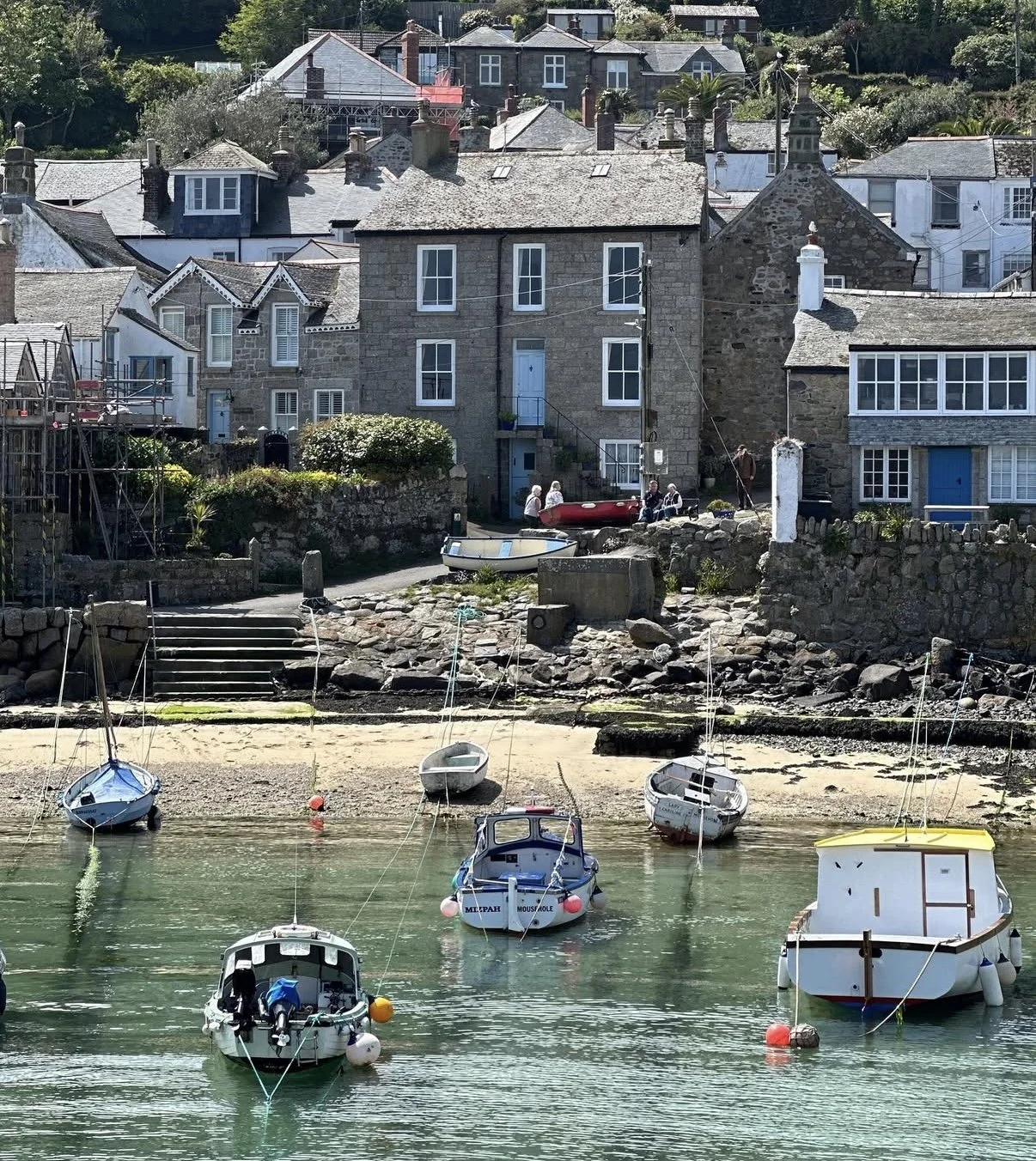 A coastal scene with several small boats anchored in calm water near a rocky shoreline. Behind the shore, there are houses built on a hillside, some with stone and wooden exteriors, and a group of people are gathered near the houses, some sitting and talking.