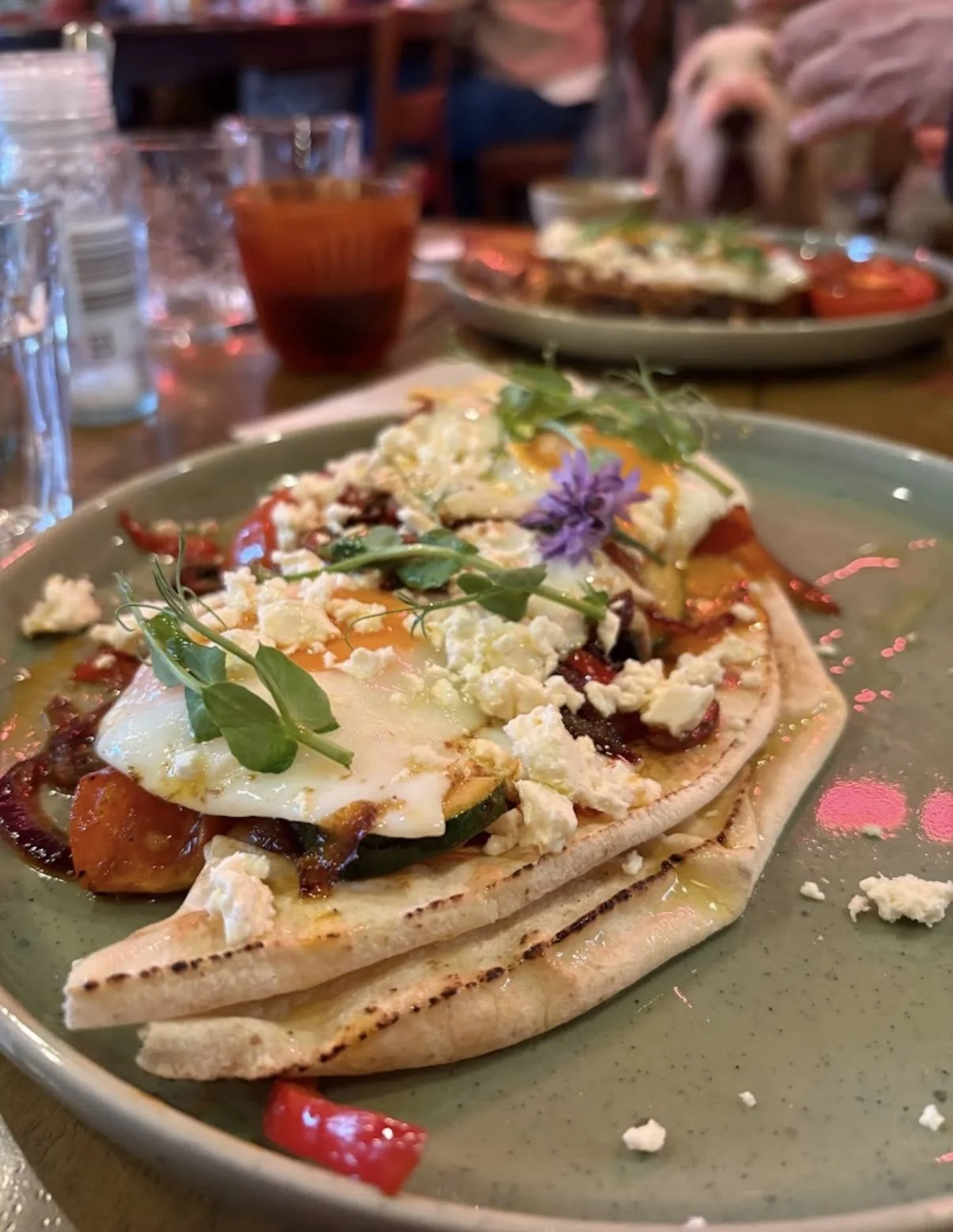 A plate of food featuring a toasted bread topped with fried eggs, crumbled cheese, roasted vegetables, and fresh herbs, garnished with edible flowers, set on a green plate in a restaurant.