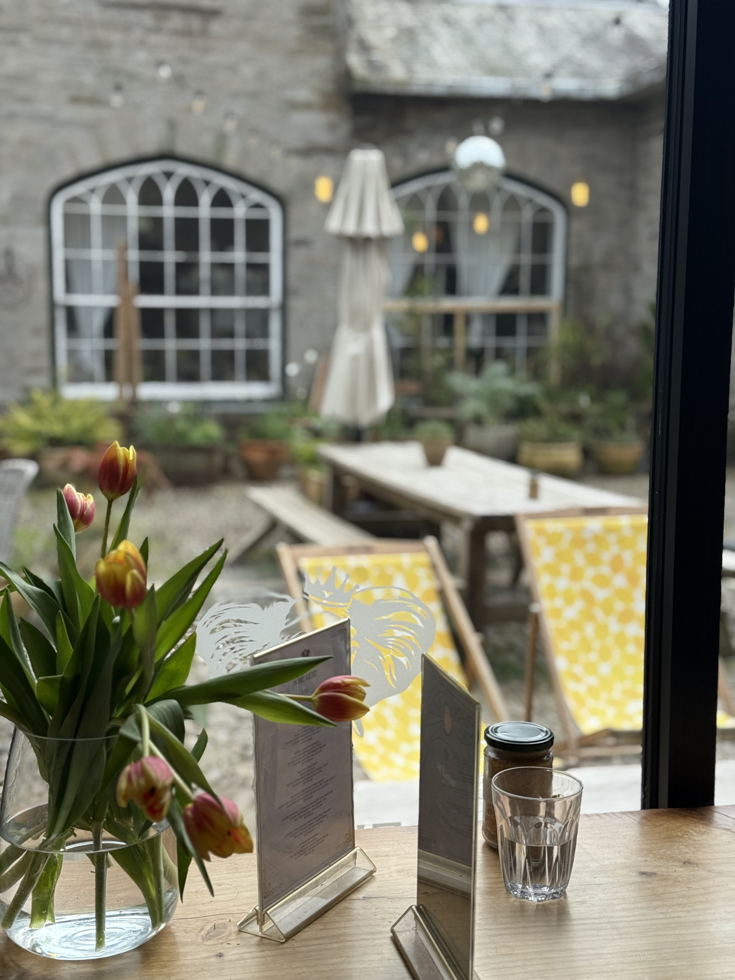 A view through a restaurant or cafe window showing a wooden table with pink tulips in a glass vase, menu cards, a glass of water, and a jar. Outside, there is a patio with yellow and white chairs, a table, an umbrella, potted plants, and a building w