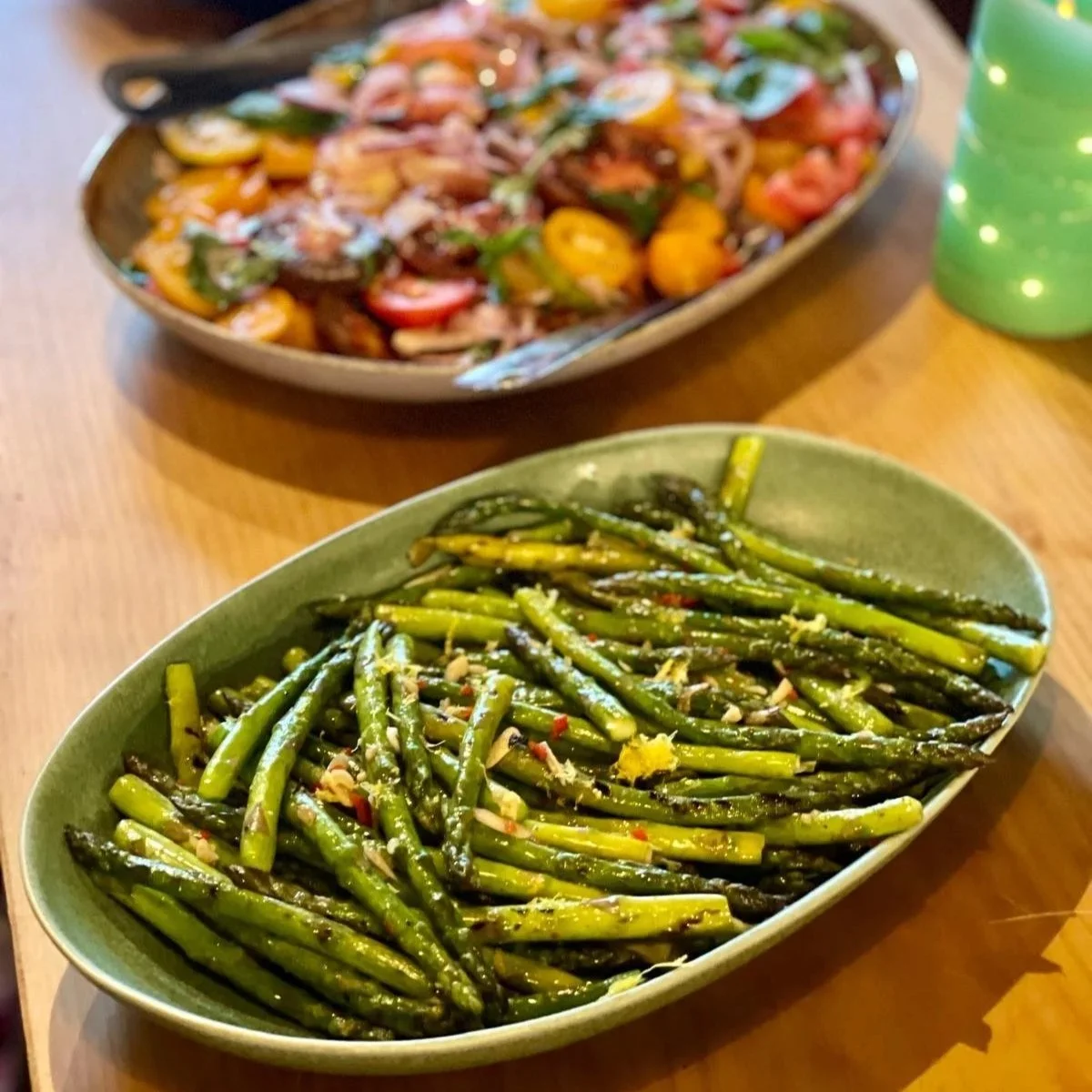 A green oval dish of cooked asparagus spears topped with garlic and red pepper flakes and a large serving bowl of a tomato and vegetable salad with cherry tomatoes, cucumbers, and herbs in the background.