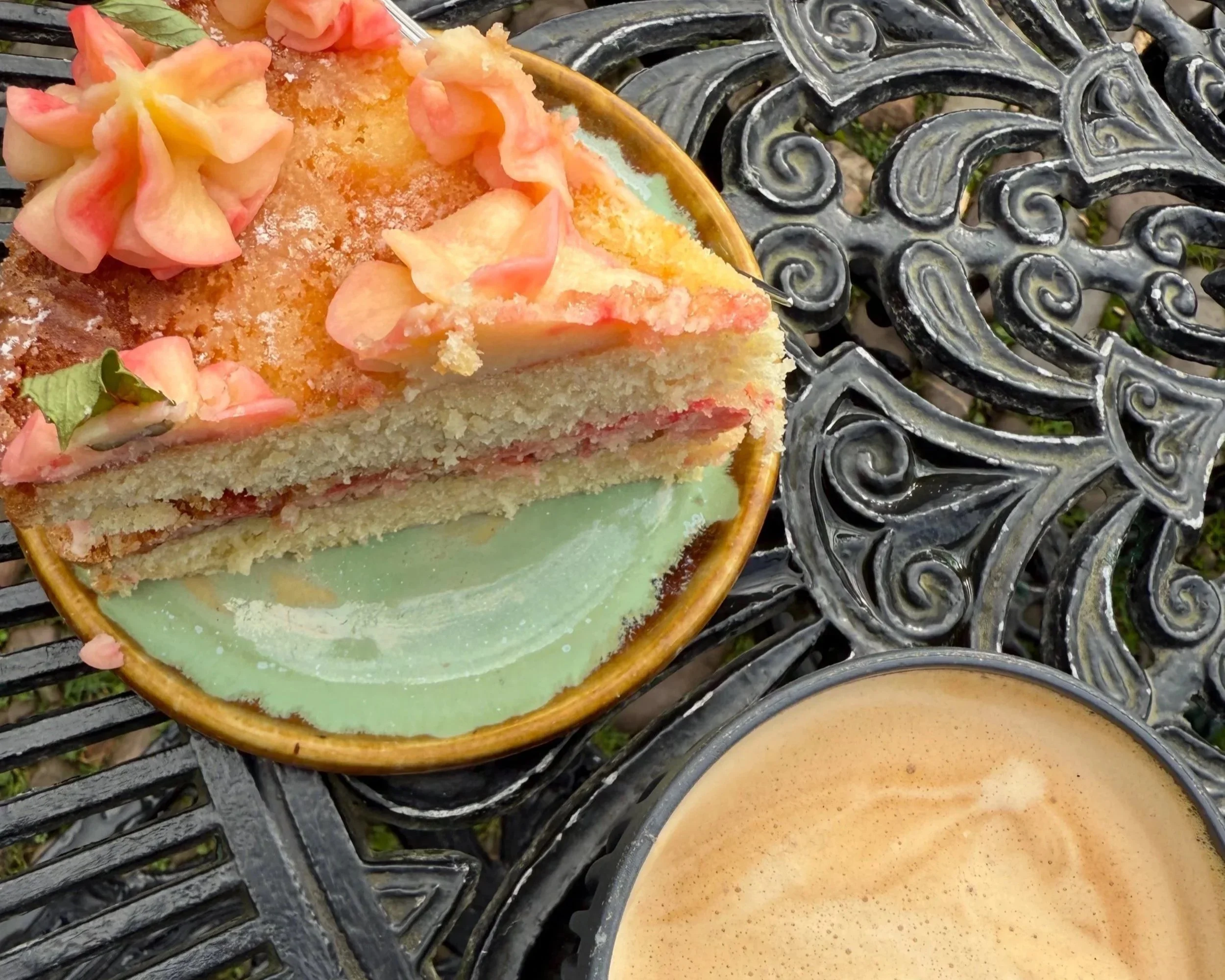 Slice of vanilla cake with pink flower decorations and green leaves, served on a light green plate, placed on a black ornate metal outdoor table.