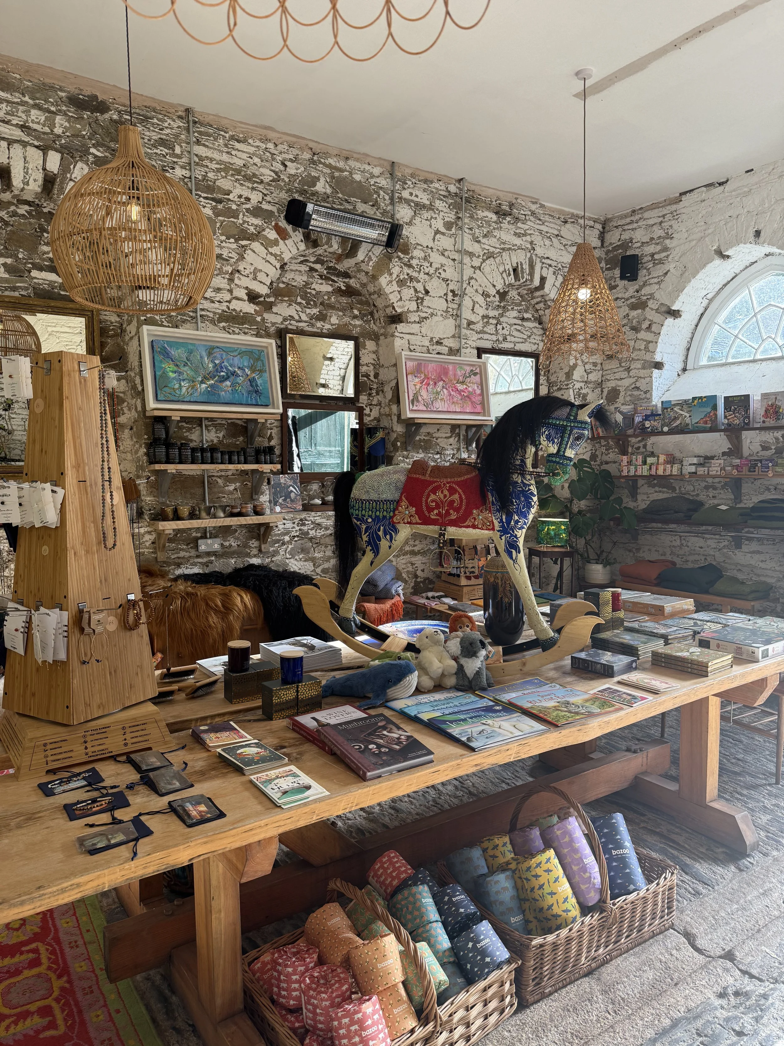 Inside a store with a rustic stone wall, displaying various items on tables and shelves, including books, toys, jewelry, and colorful wrapped textiles.