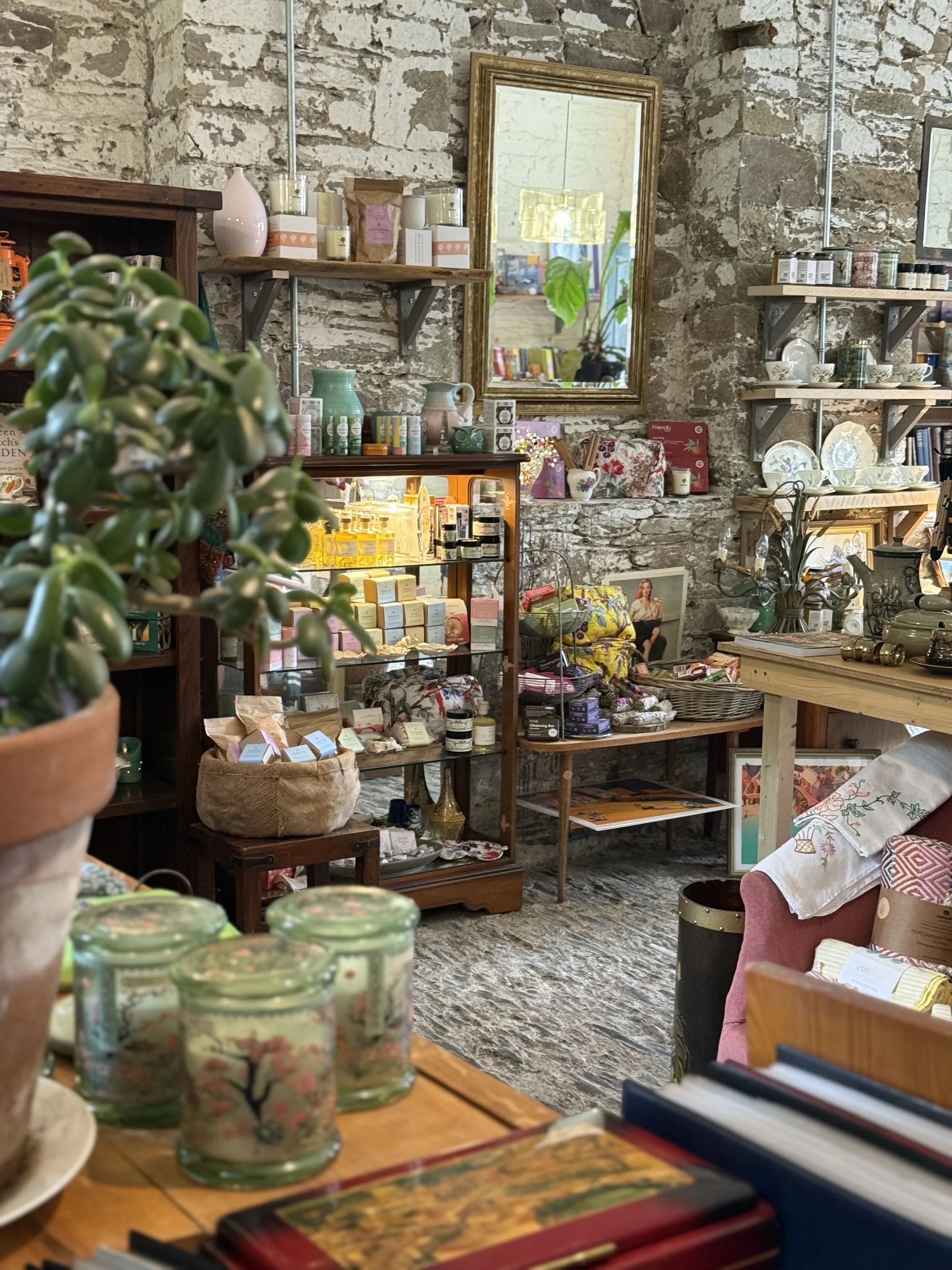 Interior of a boutique store with shelves of home decor items, jewelry, and ceramics against a stone wall, with plants and decorative objects visible.