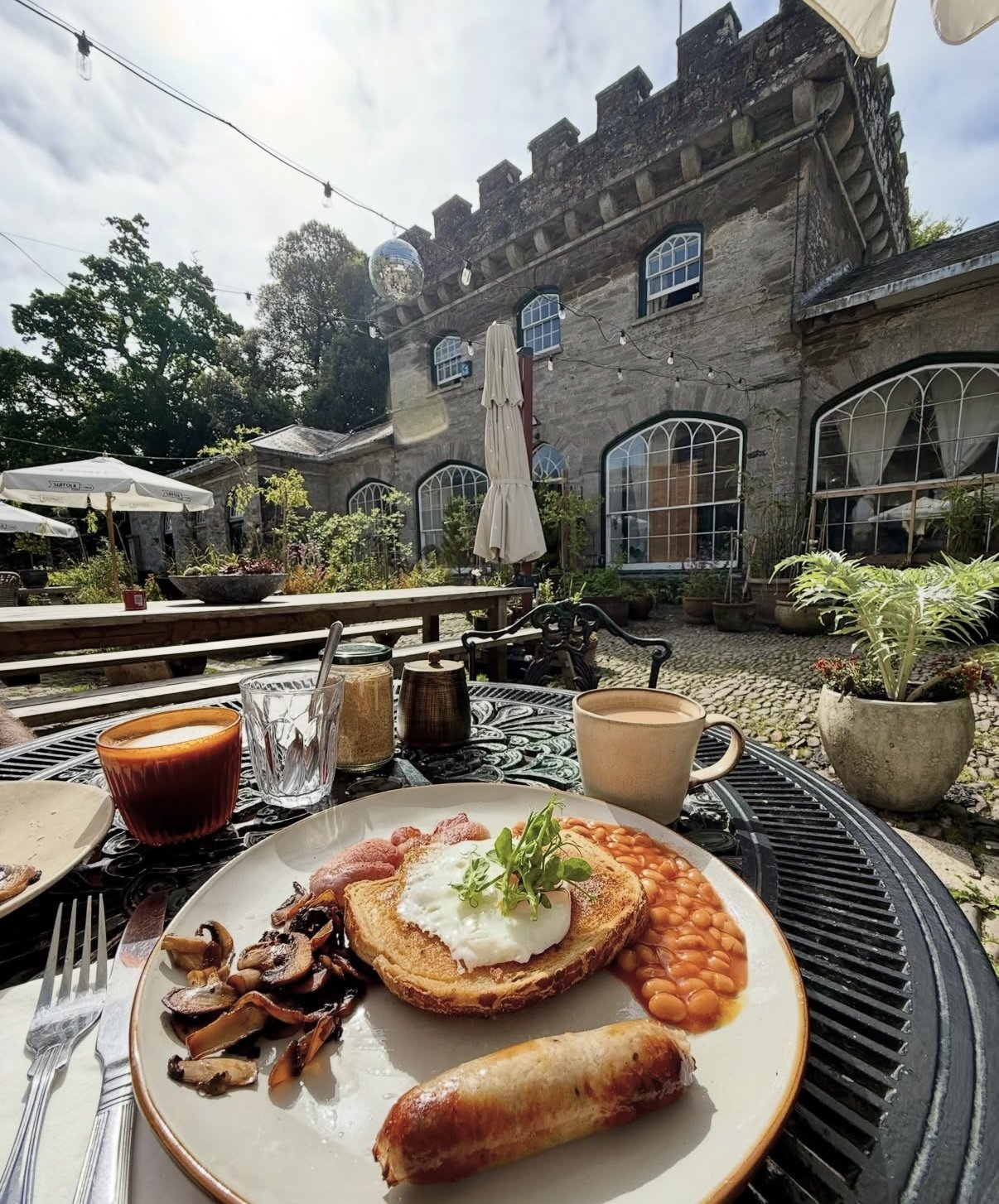 Outdoor cafe table with breakfast including eggs benedict, sausage, mushrooms, baked beans, and drinks set against a historic stone building with large arched windows and string lights in the courtyard.