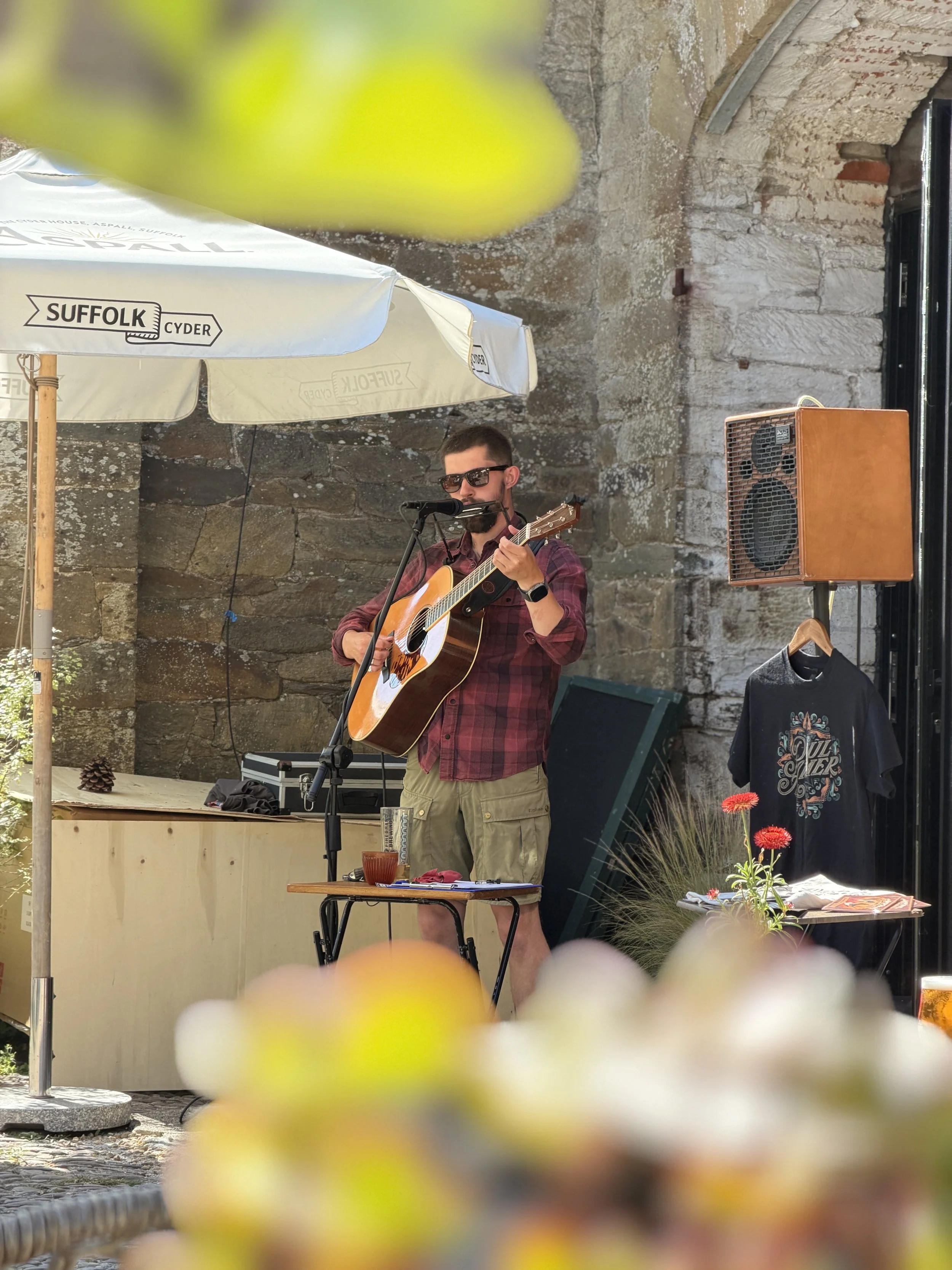 A man playing guitar and singing into a microphone outdoors under a large white umbrella with 'Suffolk Cyder' logo, with a stone wall in the background, and a table with a drink and flowers in the foreground.