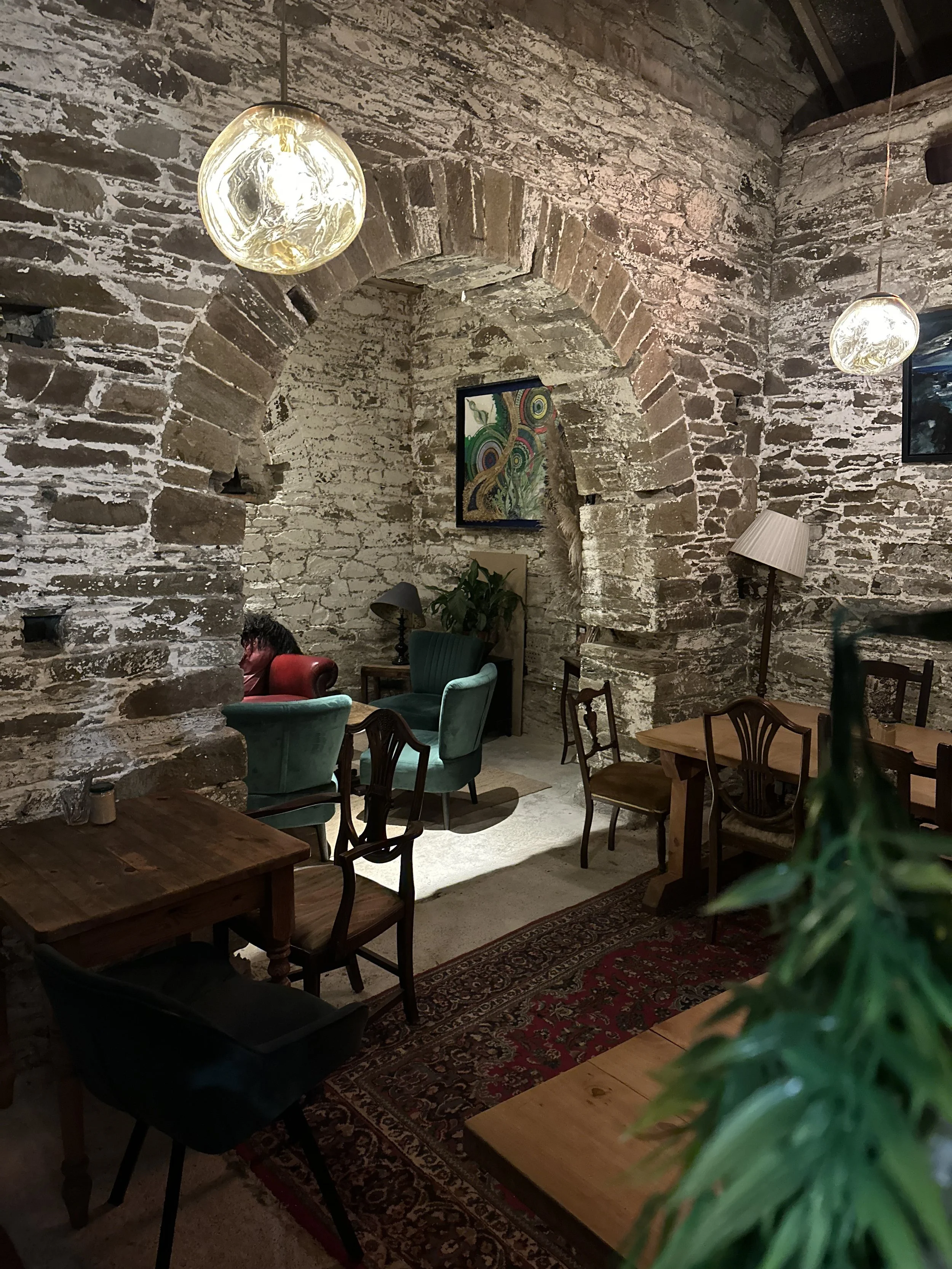 Interior of a cozy restaurant or cafe with exposed brick walls, various chairs and tables, hanging pendant lights, colorful artwork on the wall, and a patterned rug.
