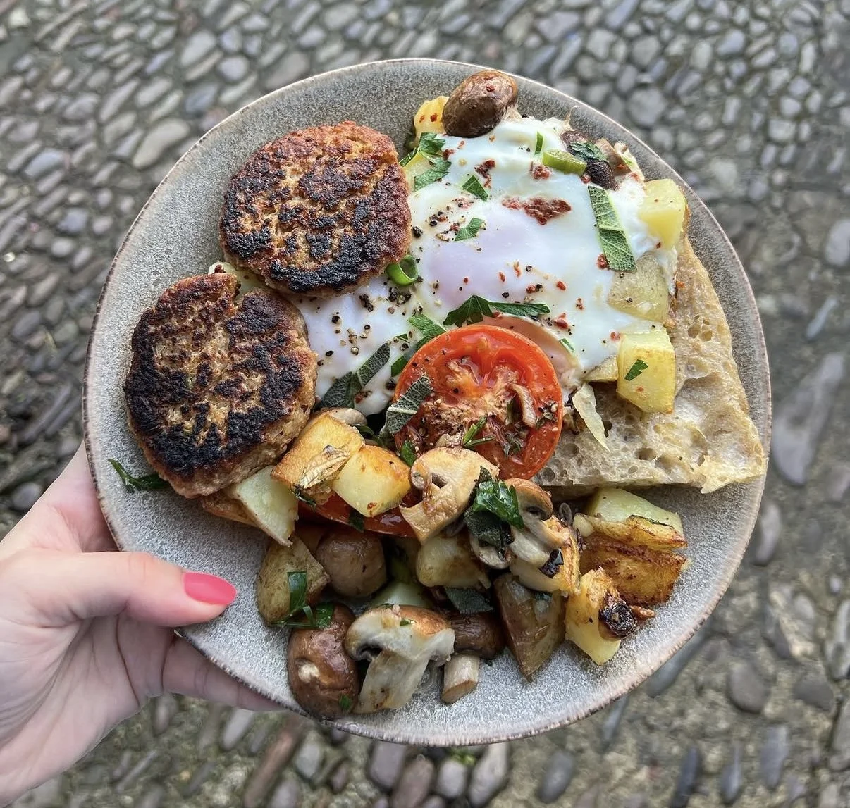 A plate of food featuring two grilled meat patties, a fried egg, a slice of tomato, sautéed mushrooms and potatoes, chopped vegetables, and garnishes, held outdoors on a cobblestone surface.