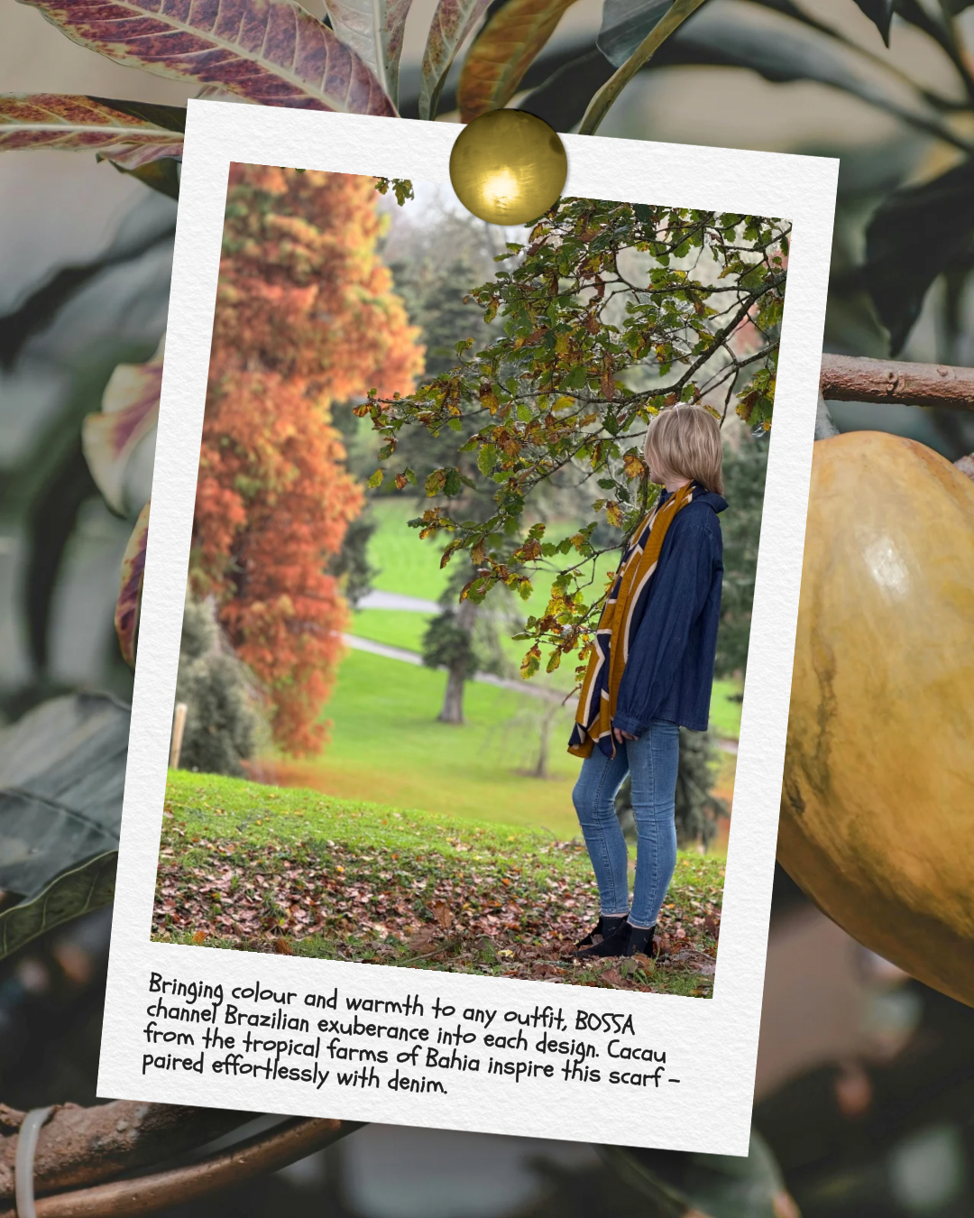 A woman standing outdoors in a park during fall, wearing denim jeans, a navy jacket, and a mustard-colored scarf, with trees and colorful autumn foliage in the background.