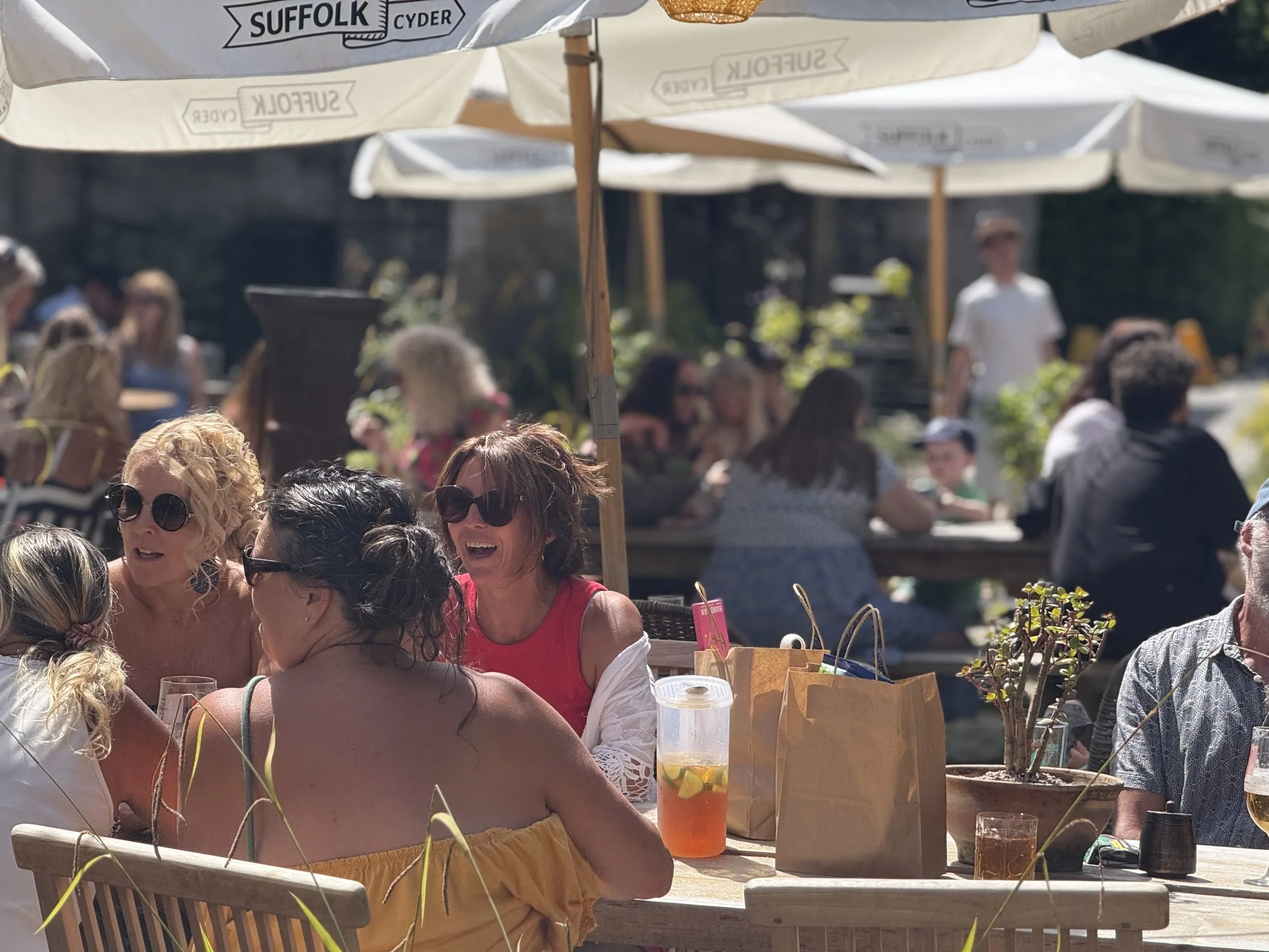 People gathered outdoors under large umbrellas, sitting at tables, enjoying drinks and conversation on a sunny day.