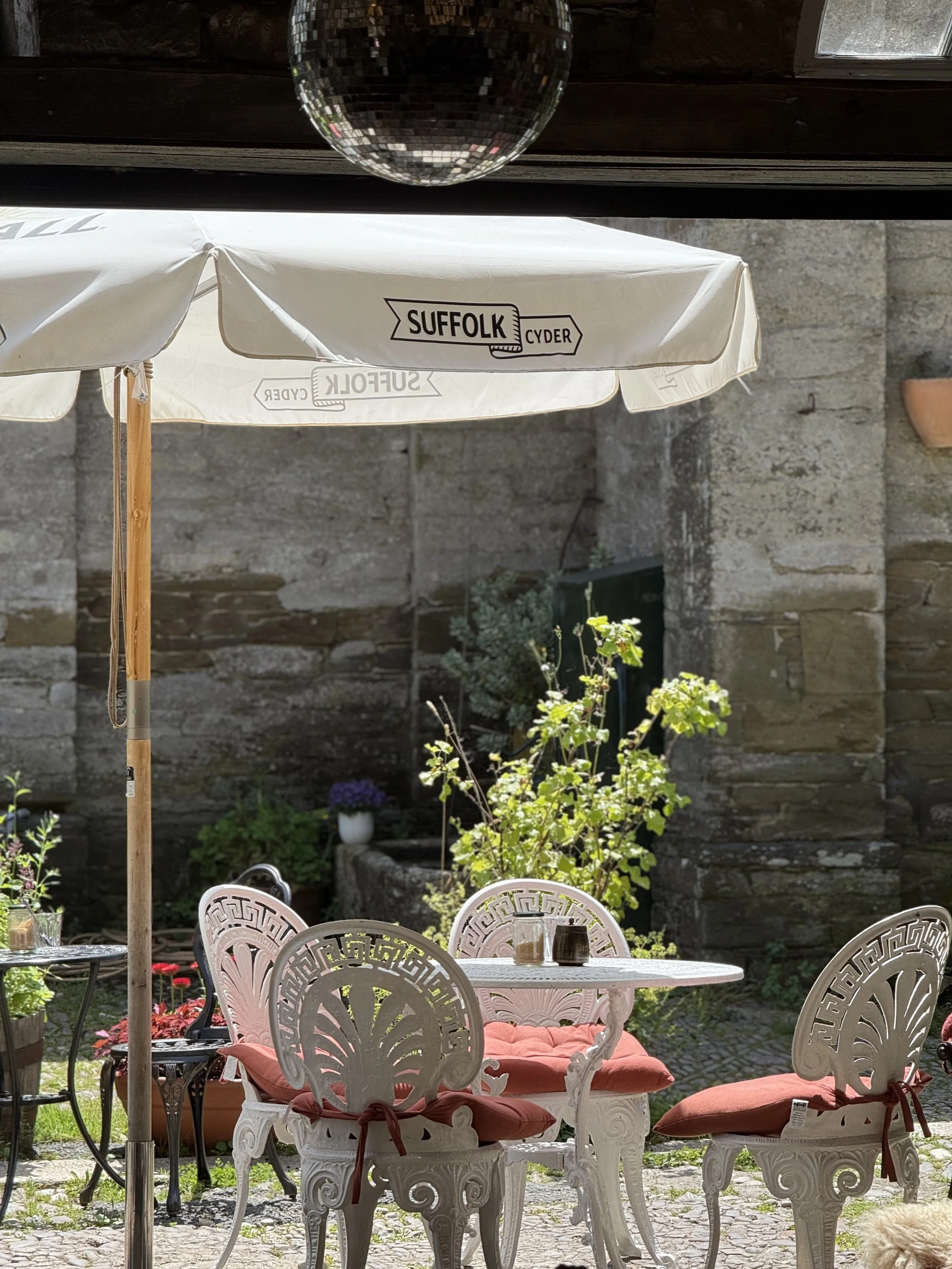 Outdoor patio scene with white ornate chairs and a table with orange cushions, surrounded by greenery and potted plants, with a stone wall in the background and a white umbrella with the words 'Suffolk Cyder'.
