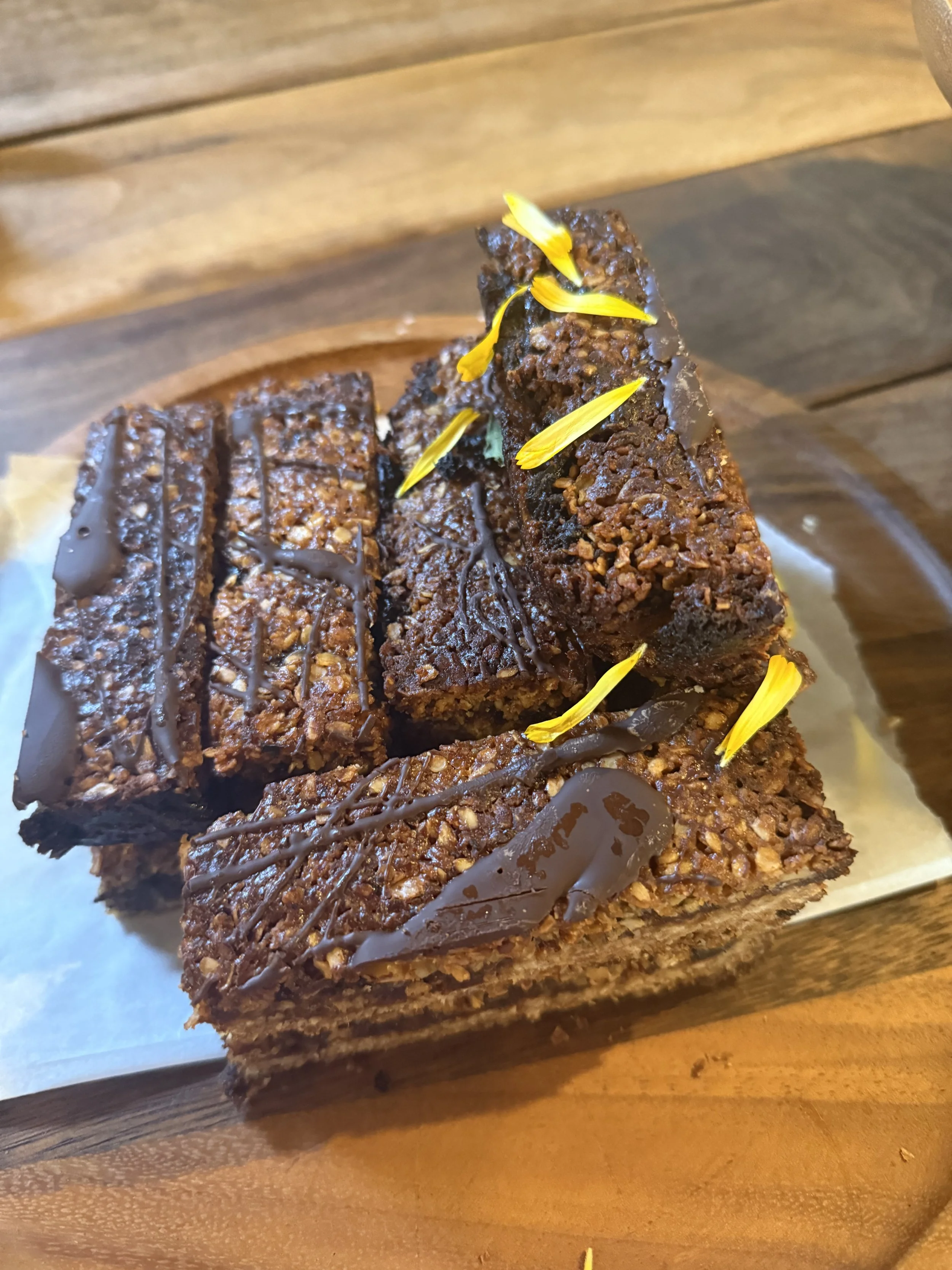 Close-up of a layered dessert bar with chocolate drizzle, topped with yellow flower petals, on a wooden tray.