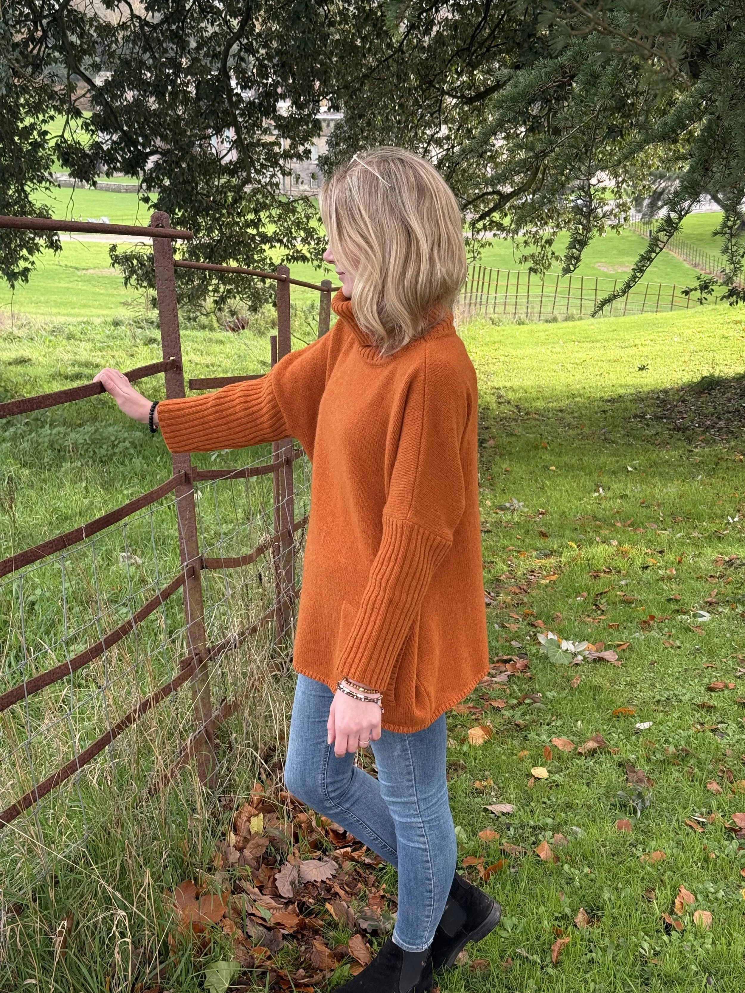 A woman with blonde hair wearing a rust-colored sweater and blue jeans stands outdoors near a rusty metal fence, holding onto it, with green grass, fallen leaves, and trees in the background.