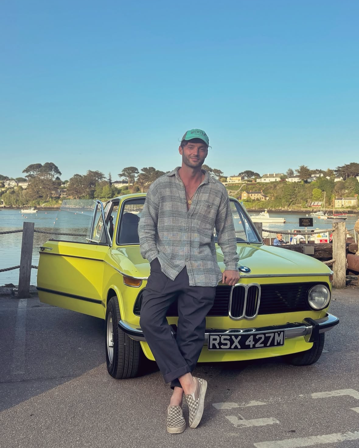 A young man leaning against a bright yellow vintage BMW car, near a waterfront with boats and houses in the background. He is wearing a green cap, a plaid shirt, dark pants, and checkerboard slip-on shoes.