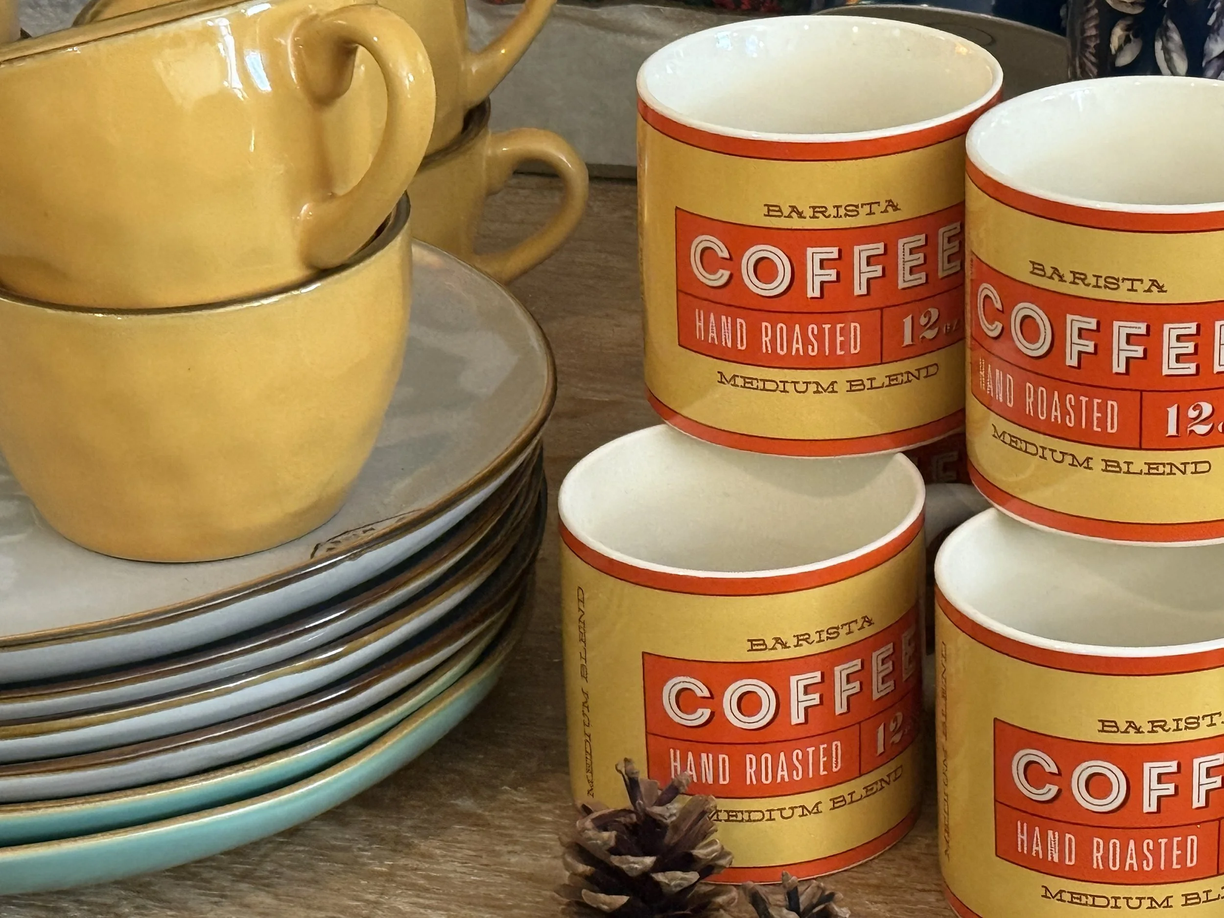 Yellow coffee mugs stacked on a plate and several yellow paper coffee cups with red and white labels that say 'Barista Coffee Hand Roasted Medium Blend', sitting on a wooden surface alongside some star anise.
