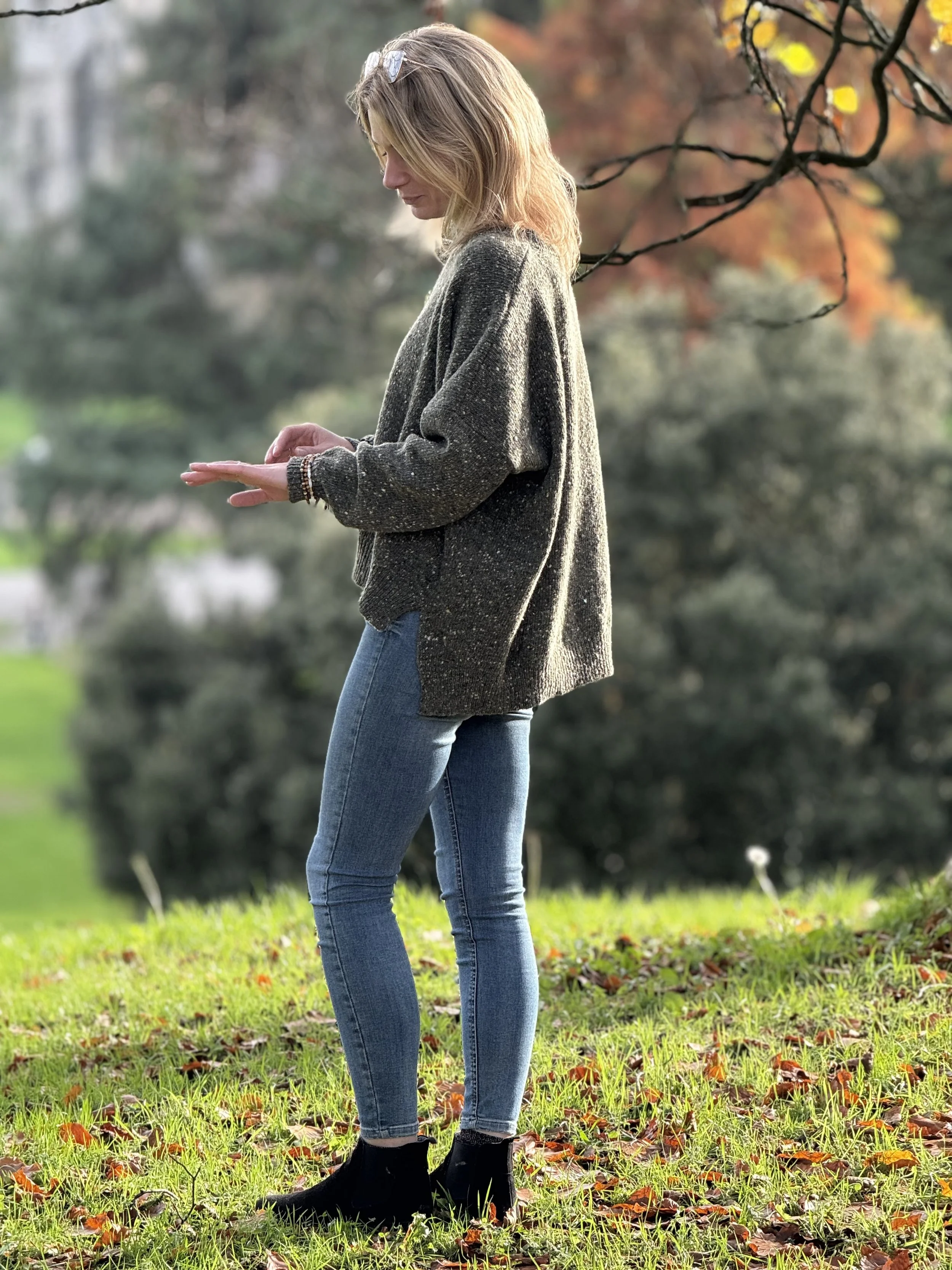 A woman with blonde hair, glasses on her head, wearing a gray sweater, jeans, and black boots, standing outdoors on grass with fallen leaves, looking at her hand. There are blurred trees with fall foliage in the background.