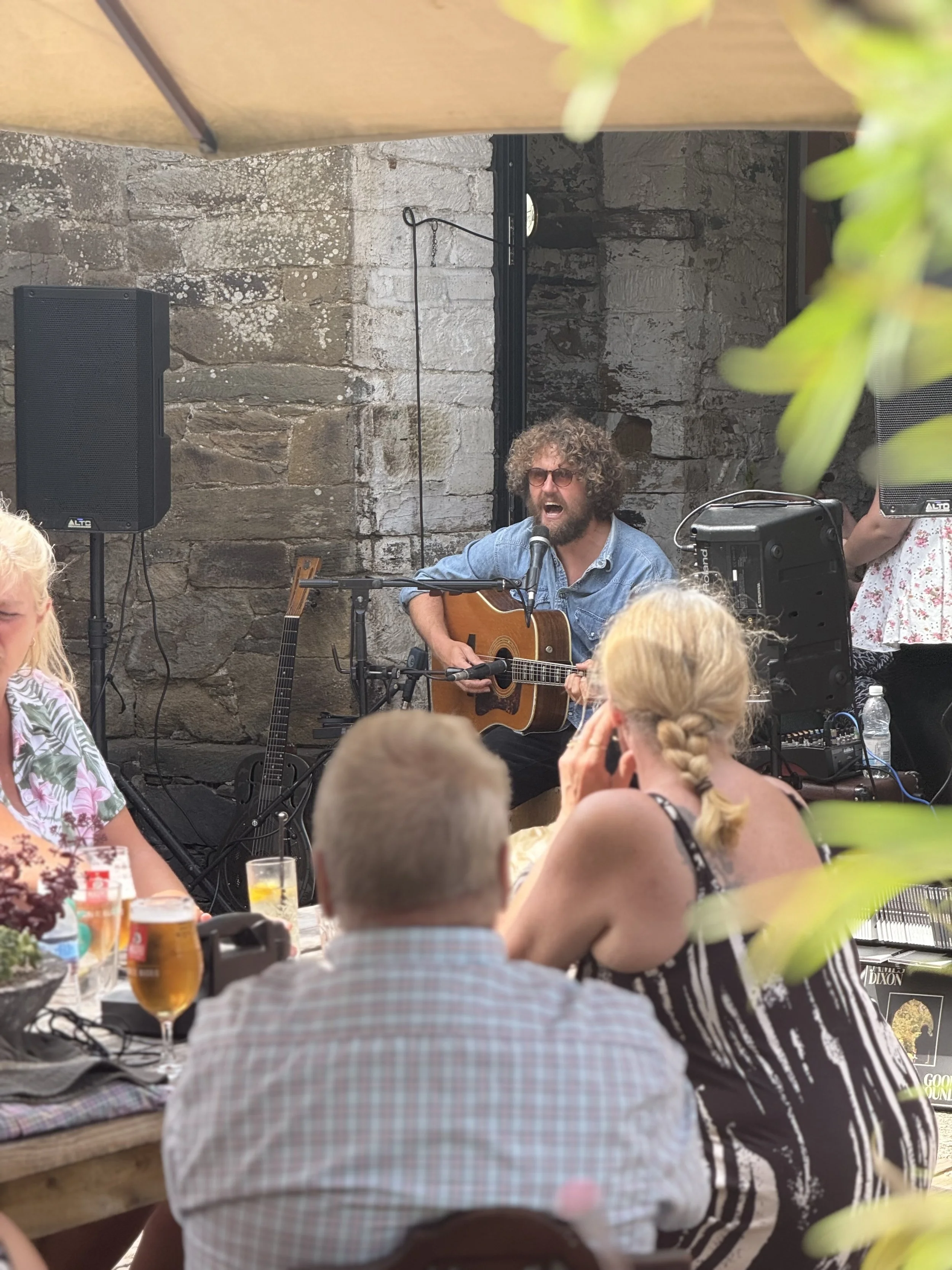 A man with curly hair, wearing sunglasses and a denim shirt, singing and playing an acoustic guitar at an outdoor event, surrounded by people dining and drinking.
