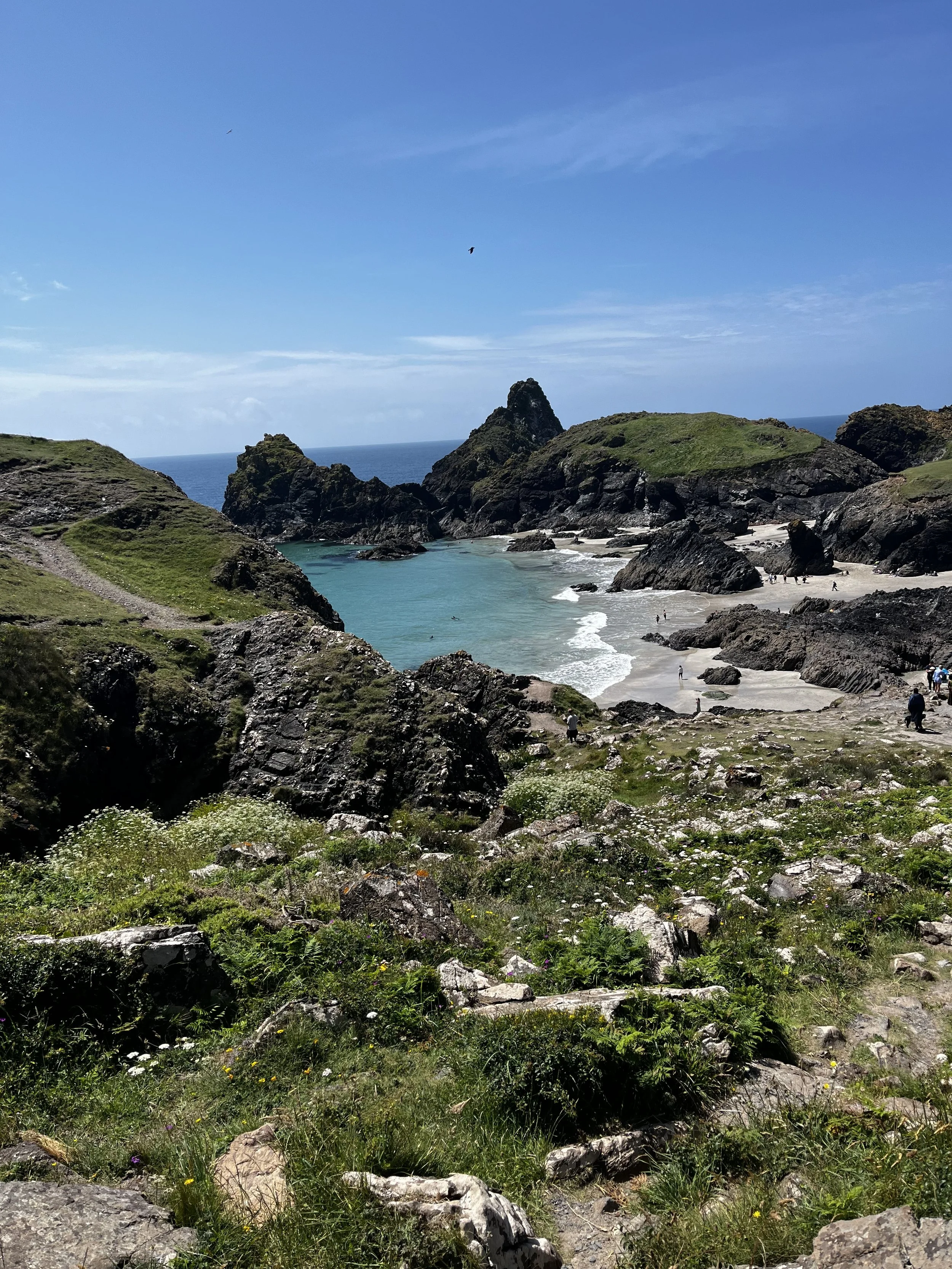 A coastal landscape with rocky cliffs, green hills, sandy beach, and ocean waves, under a partly cloudy blue sky with birds flying.