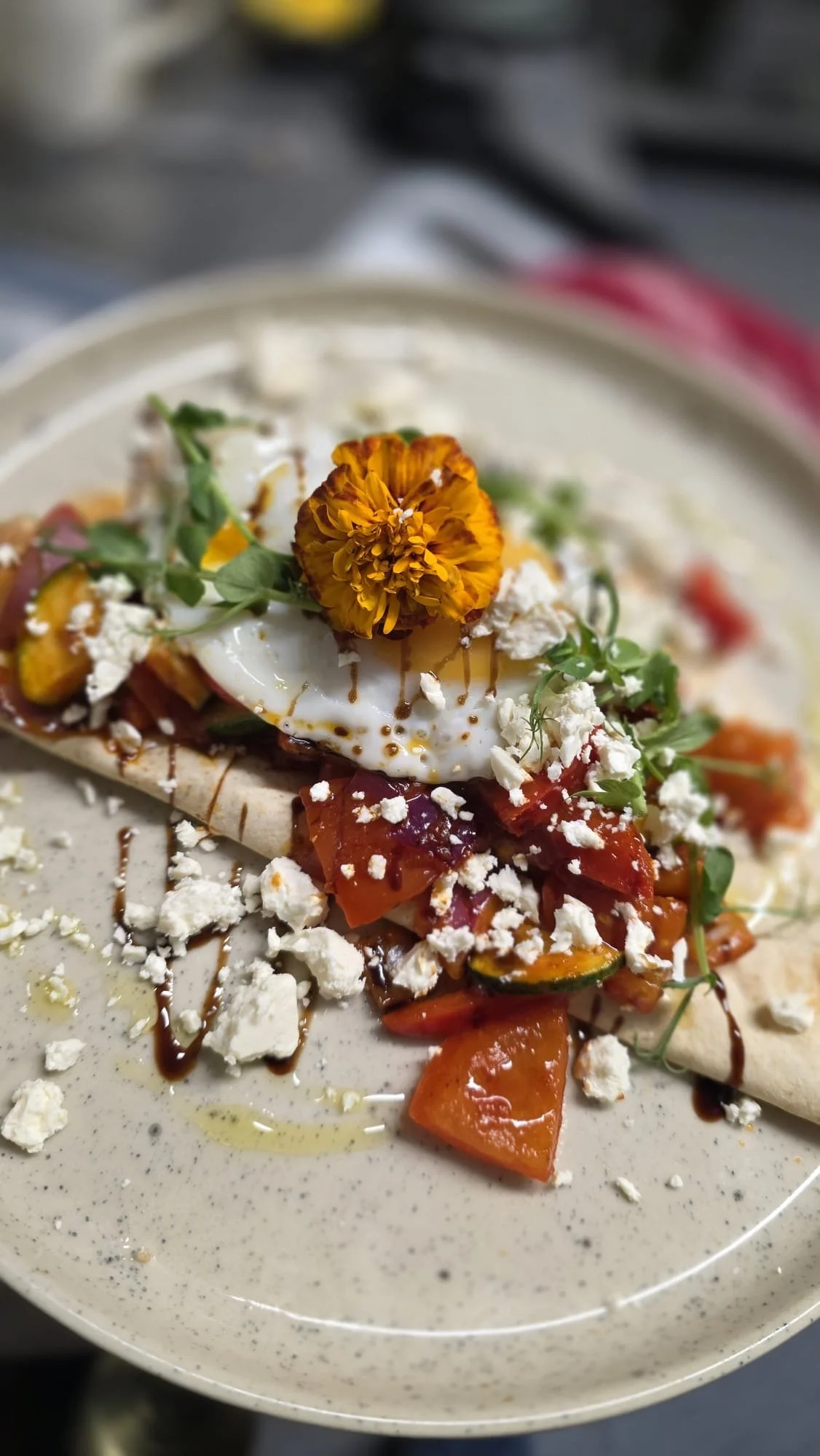 Close-up of a savory dish with a fried egg, shredded cheese, microgreens, chopped vegetables, and a flower on top, served on a beige plate.