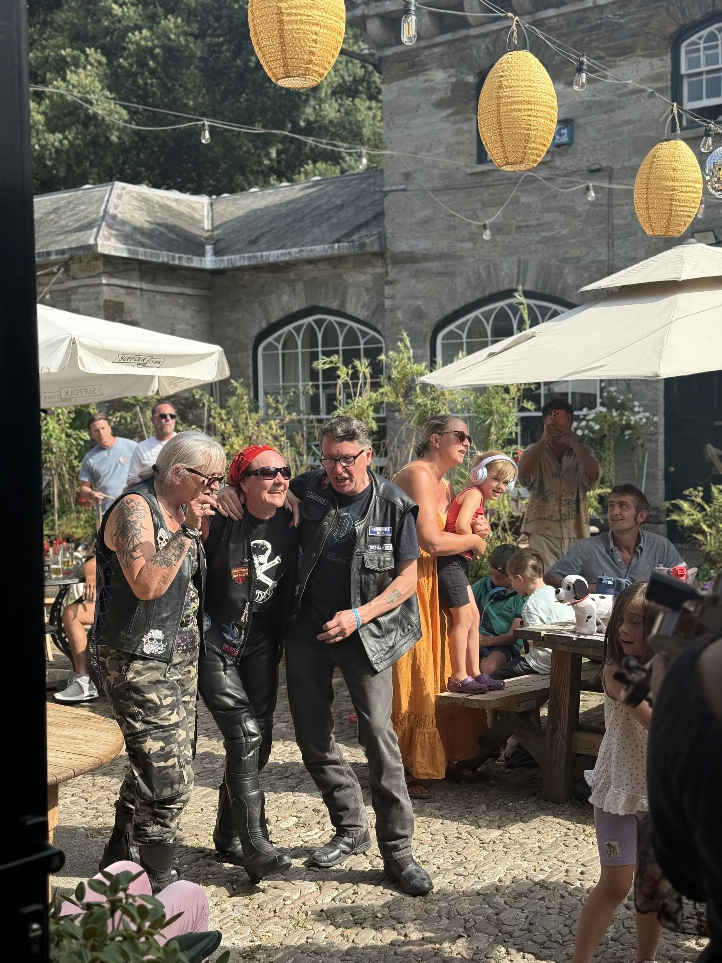 A lively outdoor gathering with people socializing under hanging yellow lanterns and string lights on a sunny day. The scene includes a stone building with arched windows in the background, some tables with umbrellas, and a diverse group of people of