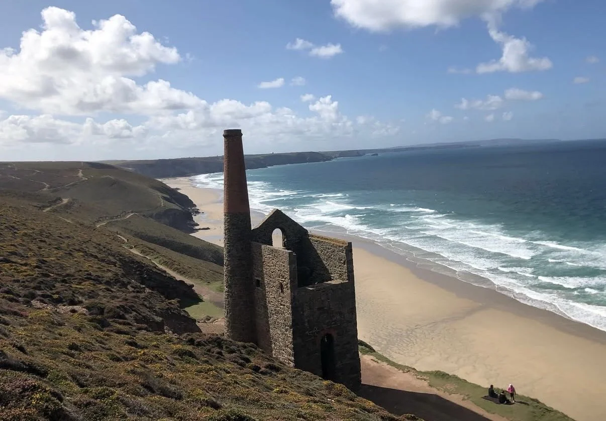 A scenic coastal landscape with an old, abandoned building with a tall chimney overlooking a sandy beach and the ocean under a partly cloudy sky.