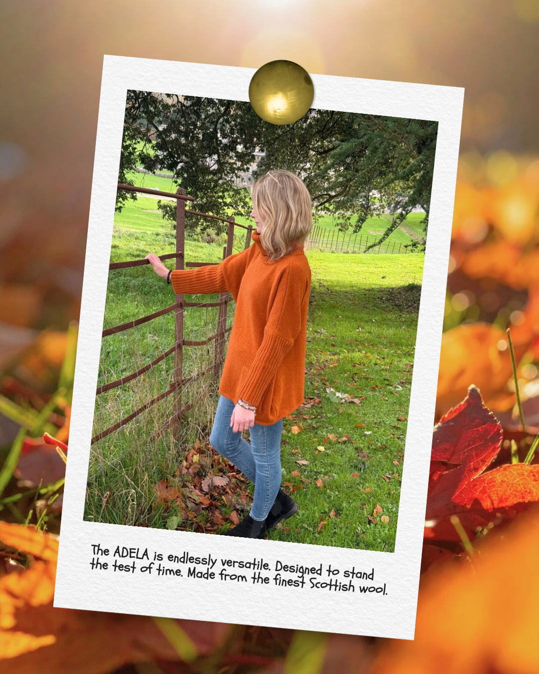 A woman wearing an orange sweater and jeans stands outdoors near a wooden fence, looking to her right with fall leaves scattered on the ground, in a green park with trees and a hill in the background.