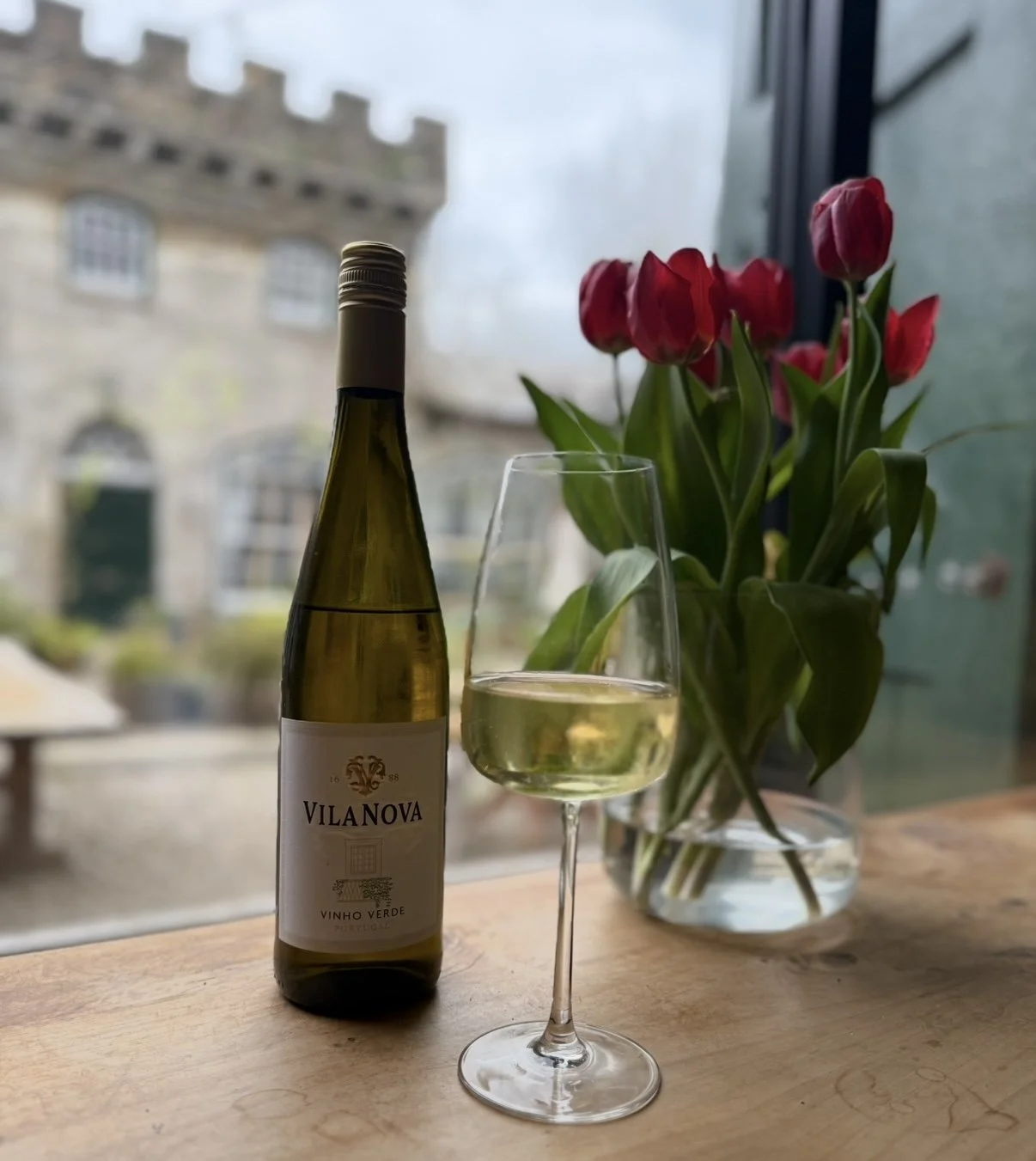 A bottle of white wine, a glass filled with white wine, and a vase of red tulips on a wooden table by a window with a view of a building outside.