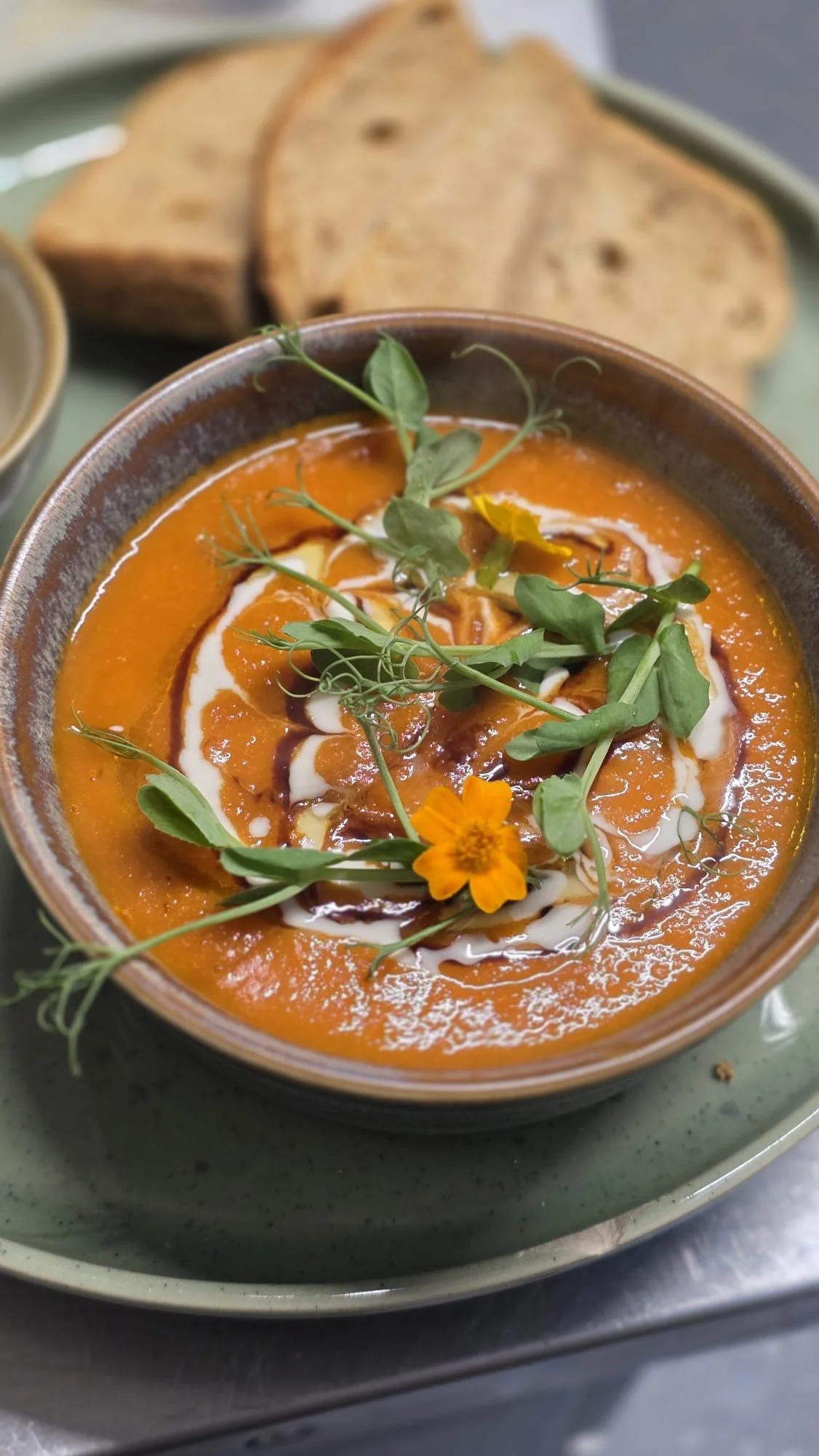 A bowl of orange-colored soup garnished with microgreens, a drizzle of cream and balsamic vinegar, with sliced bread in the background on a green plate.