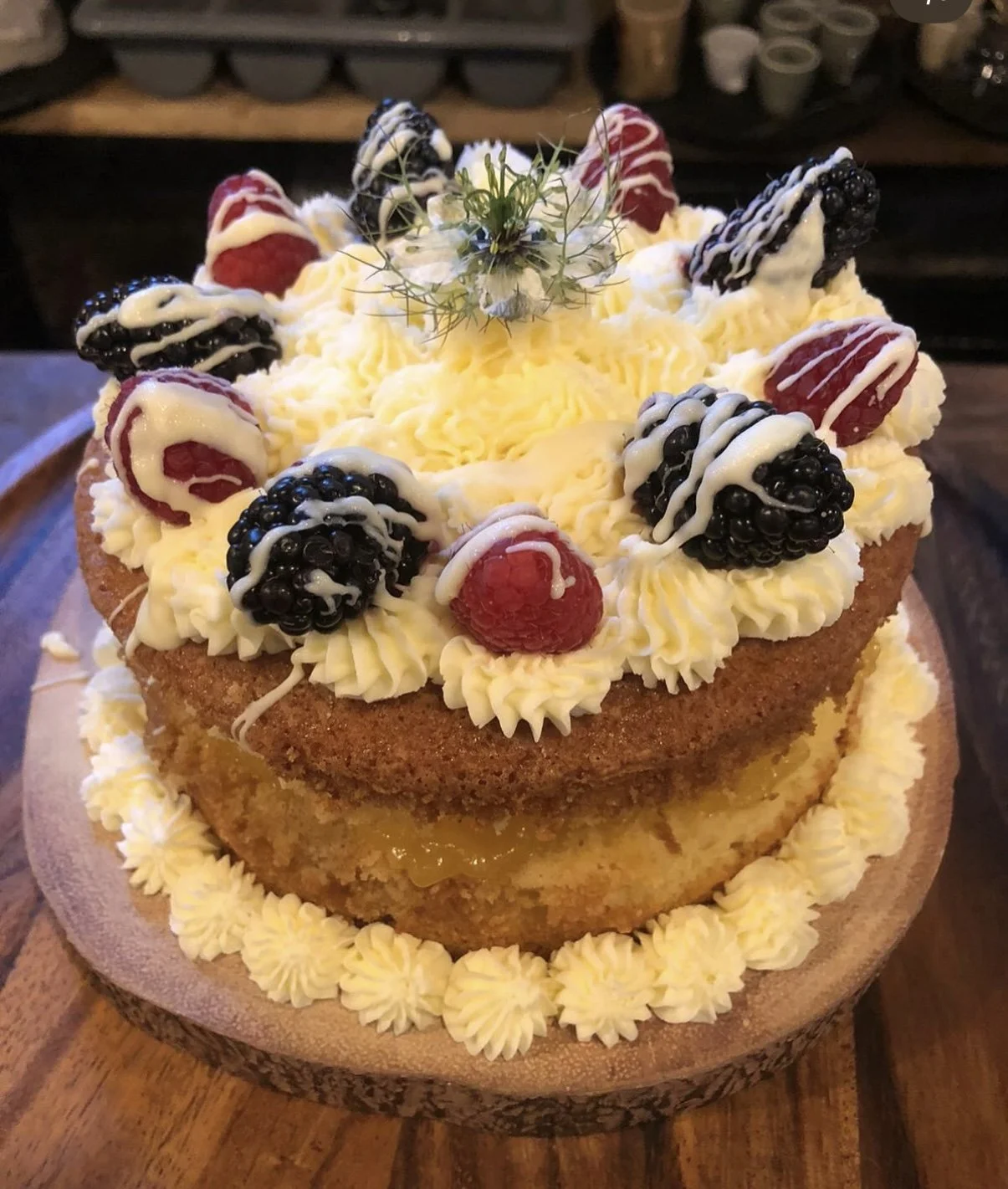 A layered cake decorated with blackberries, raspberries, and white icing, topped with a flower and herb garnish on a wooden slab.